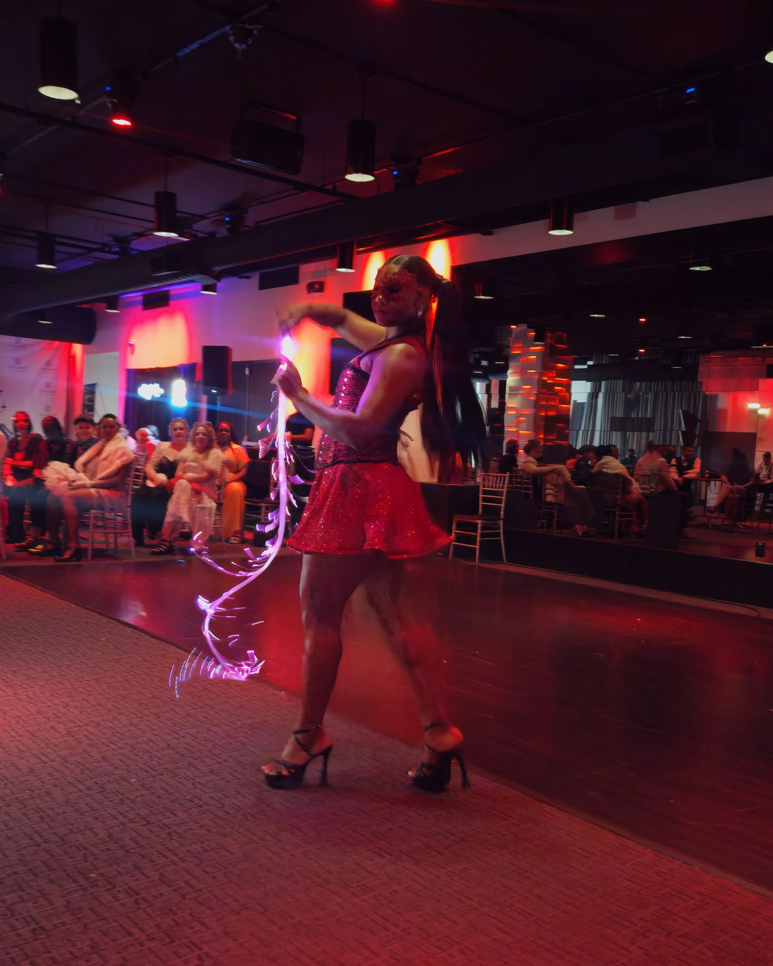 A performer wearing a red dress with sequins, high heels, and a glamorous mask performs with a ribbon in a dimly lit nightclub or dance venue. The audience watches the performance, with colorful lighting and modern decor visible in the background.