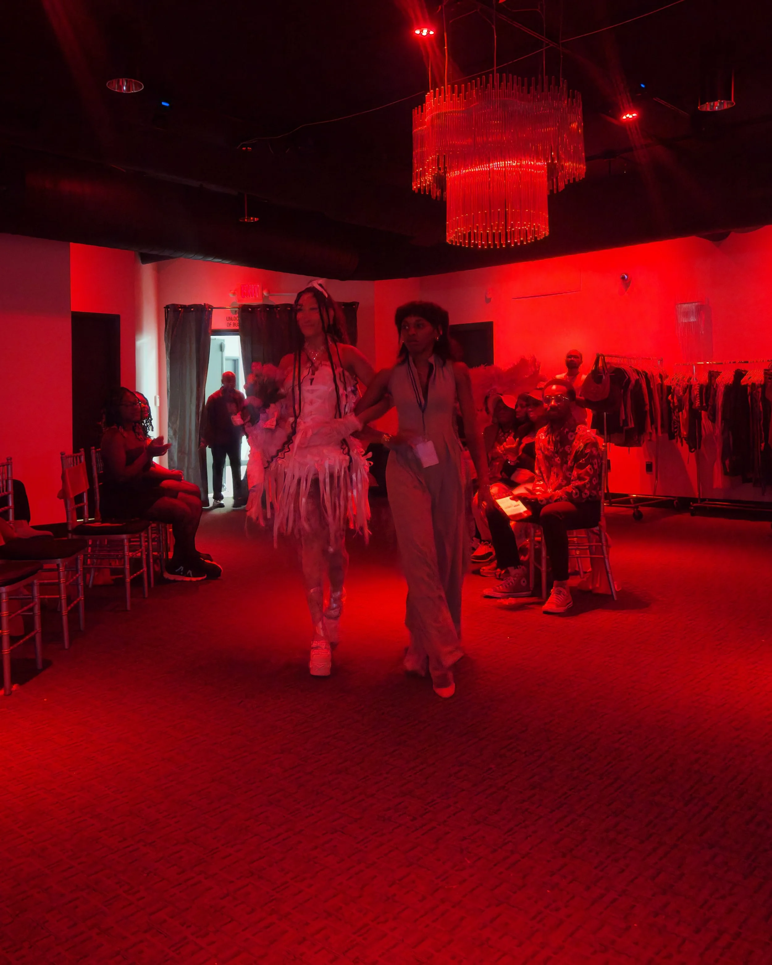 Indoor scene illuminated with red lighting, featuring two people walking together in the foreground, other seated and standing individuals in the background, and clothing racks on the right side.