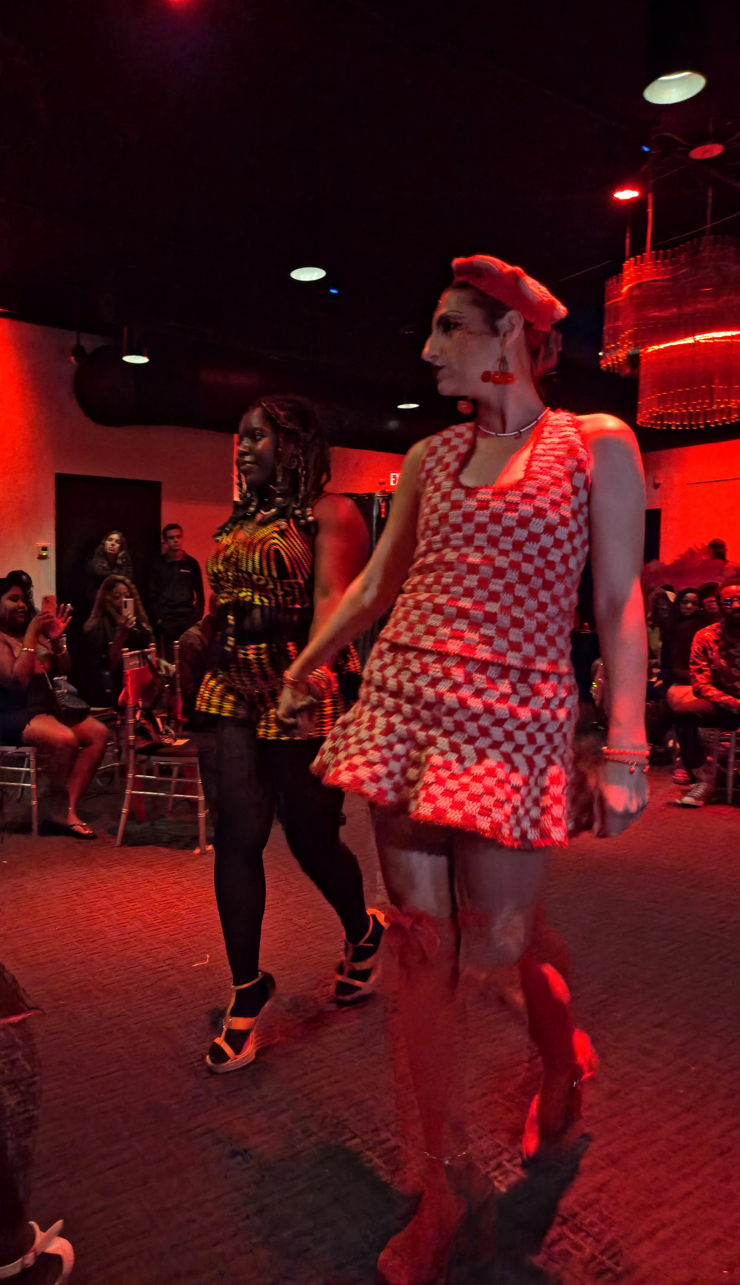 Two women are walking in a dimly lit room with red lighting. The woman in the foreground is dressed in a red and white checkered, sleeveless dress with red accessories, including earrings, a choker, bracelet, and matching red footwear. The woman in t