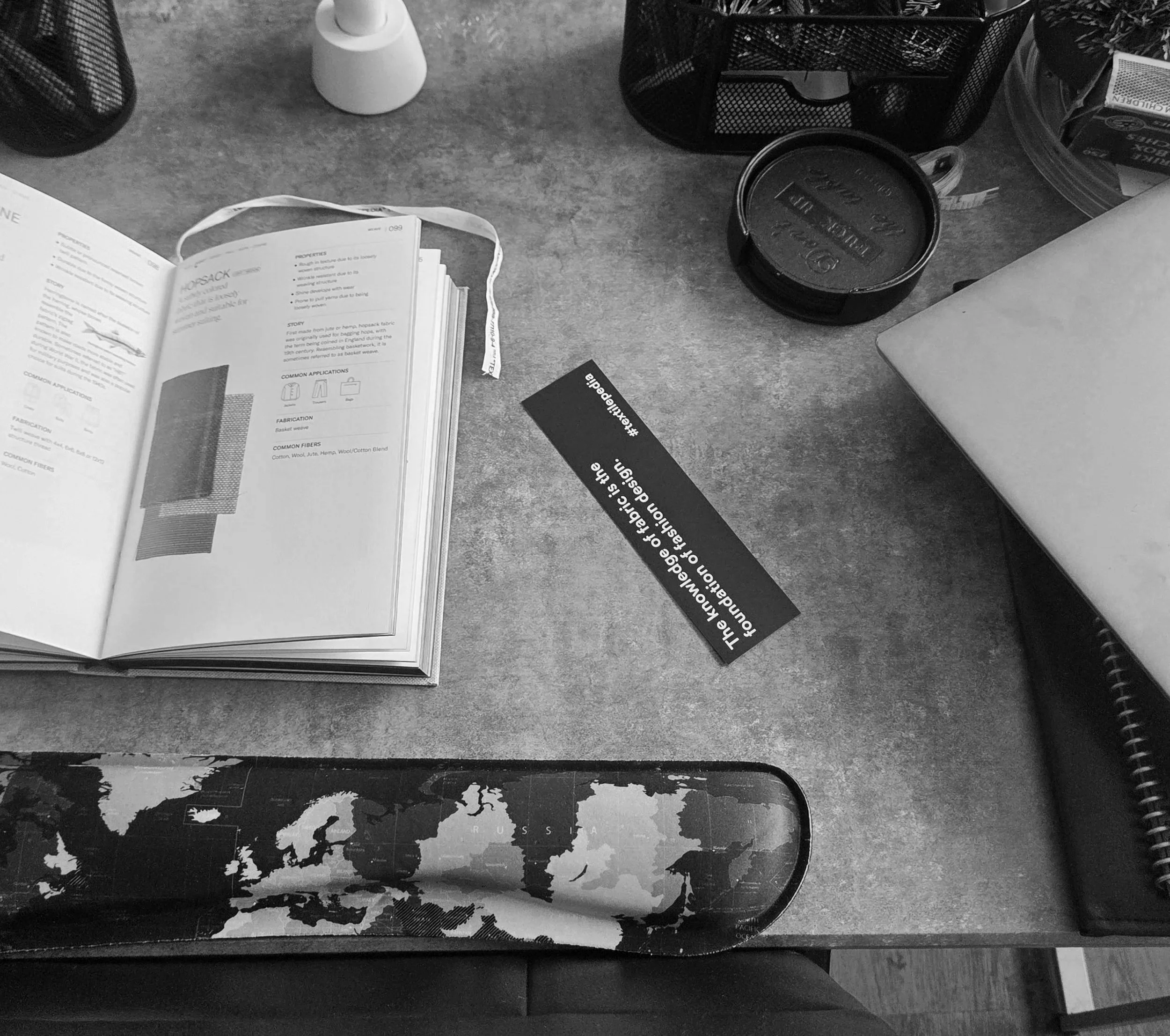 A cluttered desk with an open book, a black round container, a black mesh pen holder, a white object, and a world map on the desk surface.
