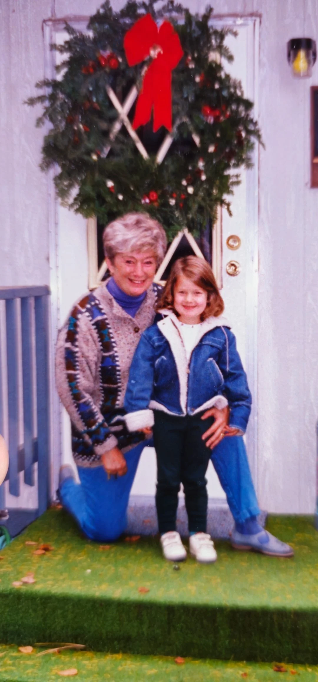 A woman and a girl standing in front of a Christmas wreath with a red bow, on a white door, in a room with white walls. The woman has short gray hair and is wearing a patterned sweater and blue jeans. The girl has shoulder-length brown hair and is wearing a blue jacket, white sneakers, and dark pants. They are smiling and posing for the photo.