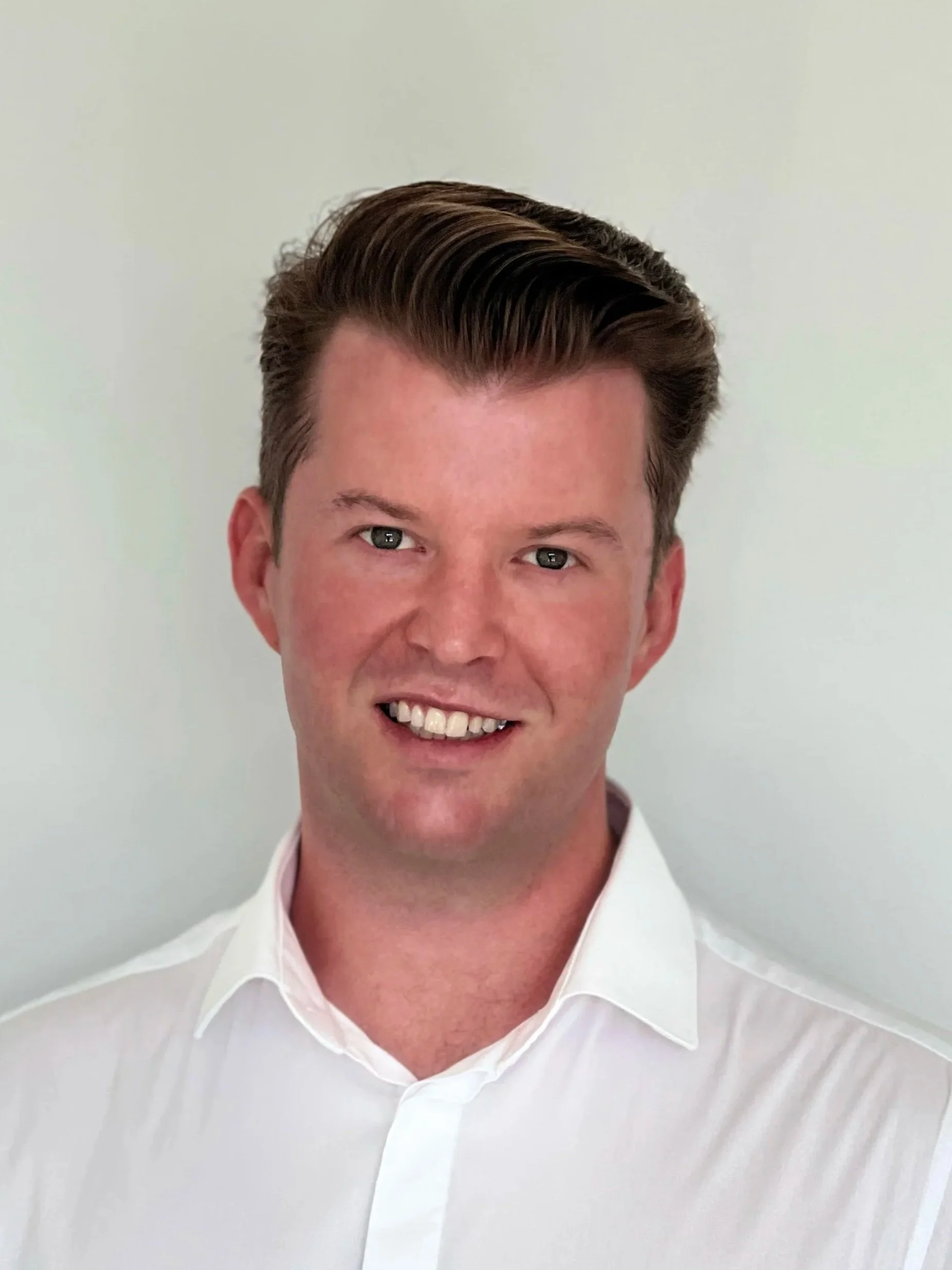Headshot of a smiling man with dark brown hair, wearing a white collared shirt, against a plain light background.