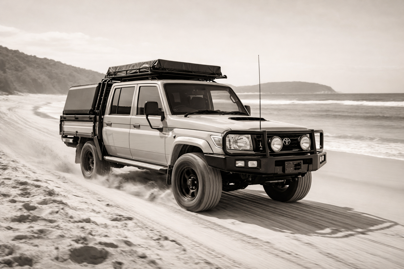 A rugged four-wheel-drive vehicle driving on a sandy beach with waves in the background.