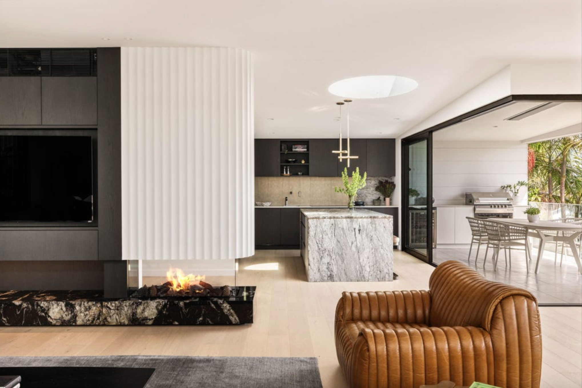 Modern open-concept living room with a fireplace, a leather armchair, and a kitchen with a marble island, pendant lights, and a sliding glass door leading to an outdoor dining area with a grill and patio furniture.