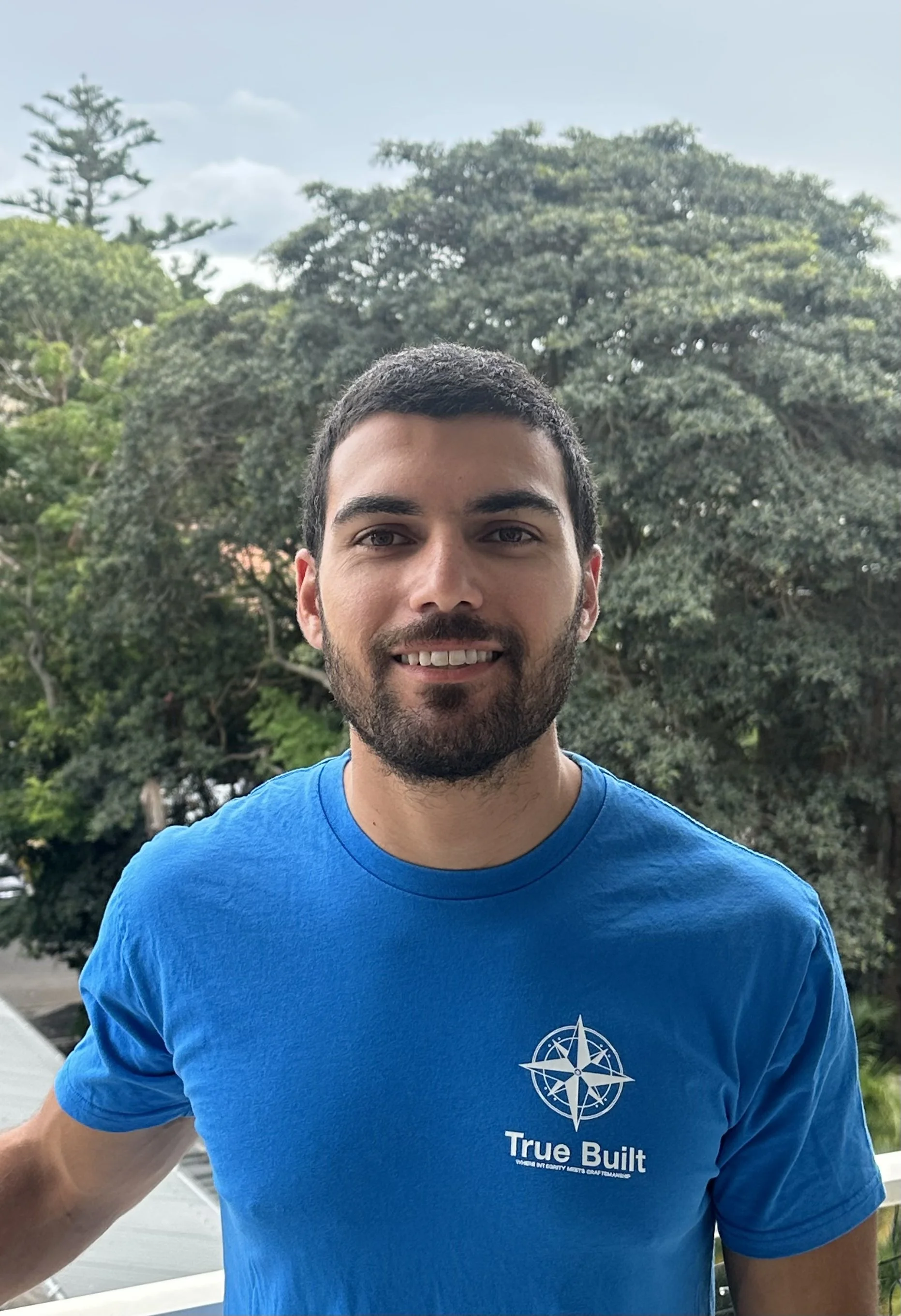 A smiling man with a beard and short dark hair wearing a blue t-shirt with 'True Built' logo, standing outdoors near trees.