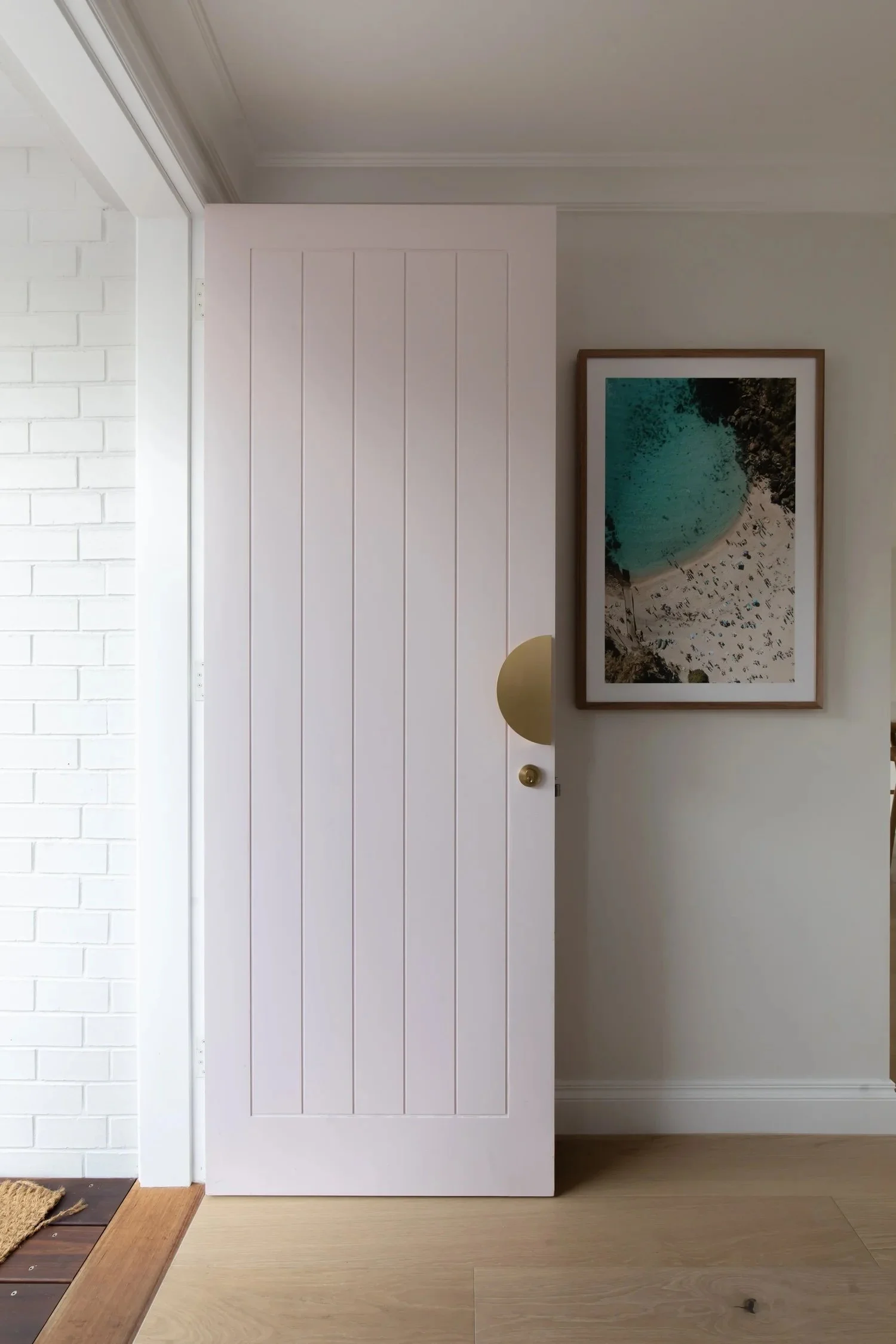 An open white door in a room with a framed aerial beach photograph on the wall. Visible parts of a white brick wall, light wood flooring, and a brass wall lamp.