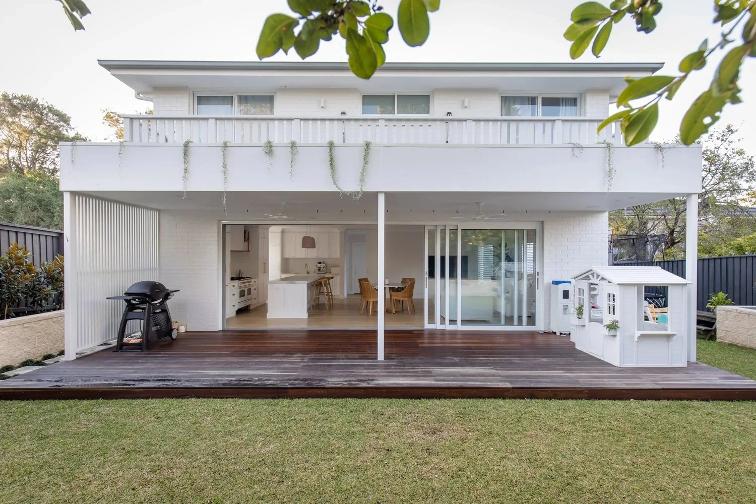 A modern two-story house with a white exterior, large sliding glass doors, an outdoor wooden deck, a barbecue grill, and a small white playhouse in the backyard.
