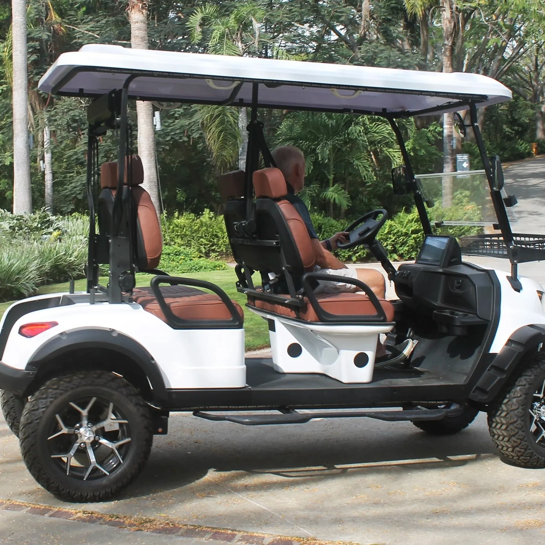A white golf cart with brown seats parked on a paved path in a tropical setting with palm trees and green shrubs, with an elderly man sitting in the driver's seat.