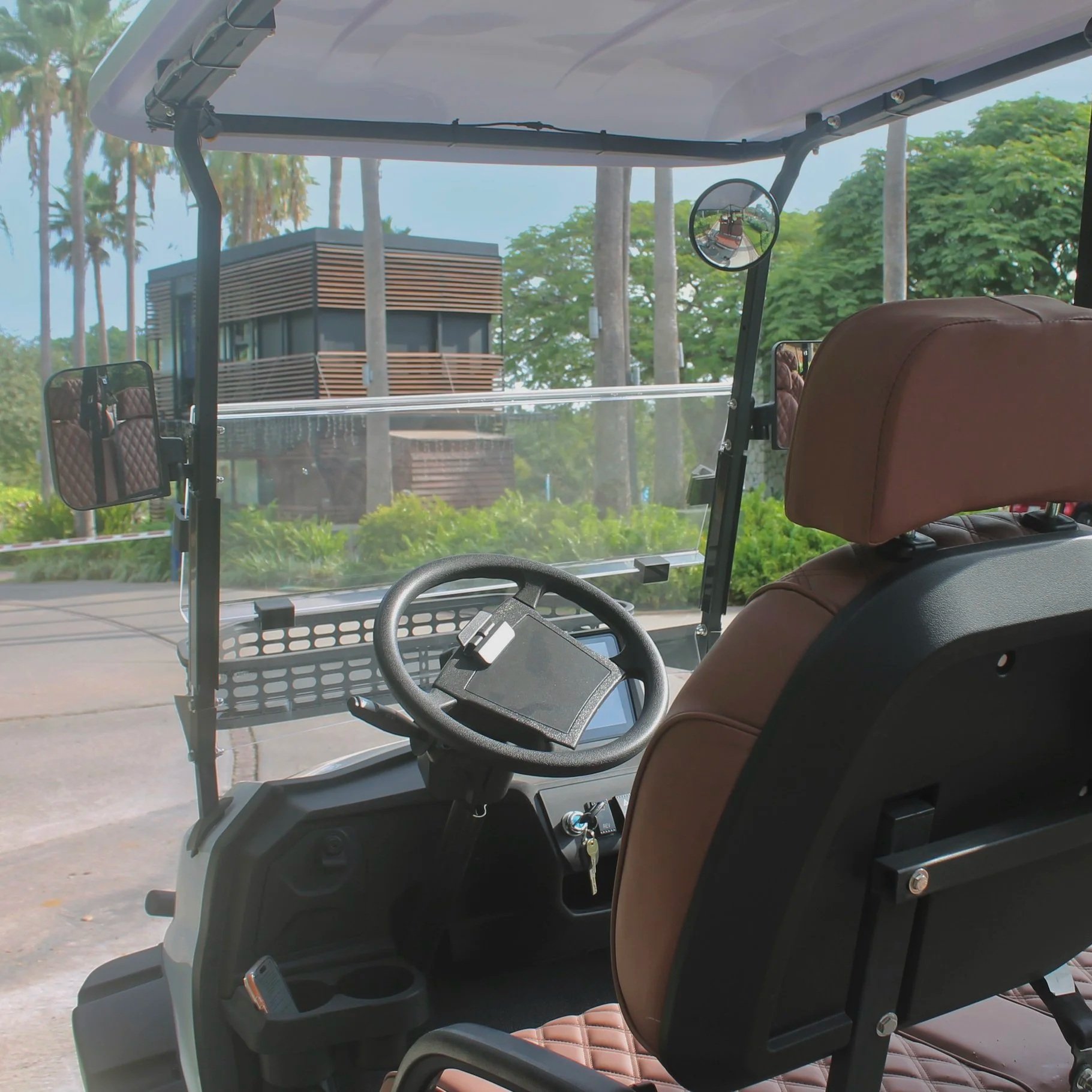 Interior of a golf cart with brown leather seats, steering wheel, rearview mirrors, and a clear windshield, with trees and a modern building visible outside.
