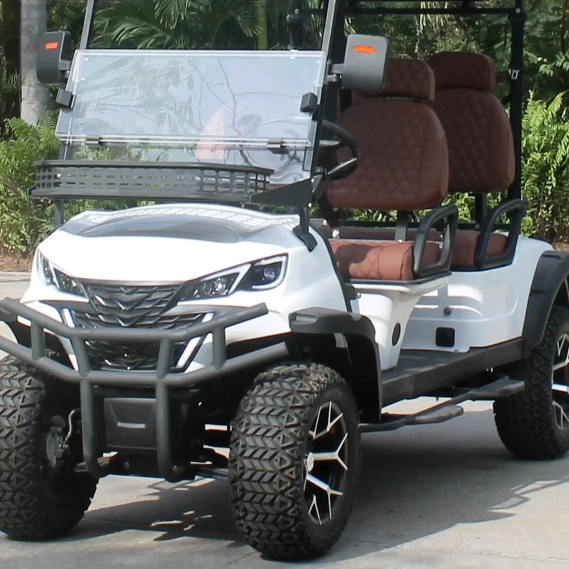 White golf cart with four brown cushioned seats, black framing, large off-road tires, and clear windshield, parked outdoors with greenery in the background.