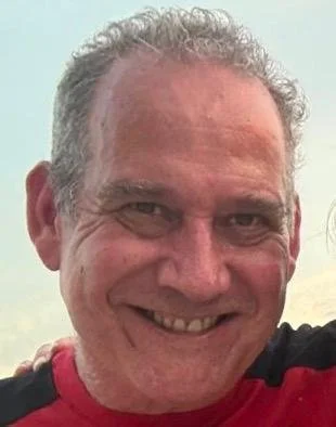 A smiling man with gray curly hair, wearing a red and black shirt, outdoors with a cloudy sky in the background.