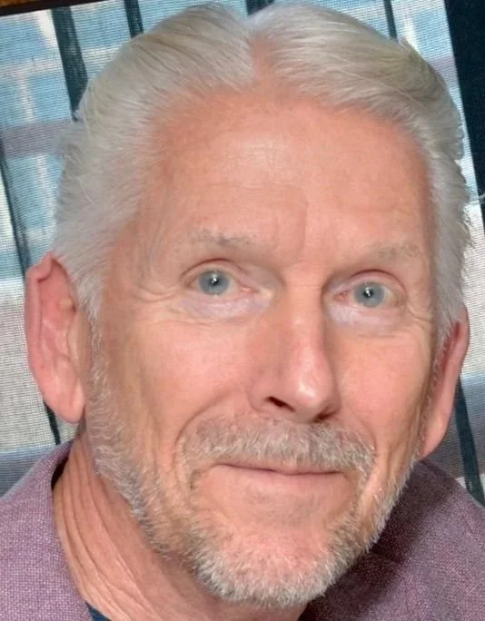 Close-up of an older man with gray hair and a beard, smiling, in front of a window with blinds.