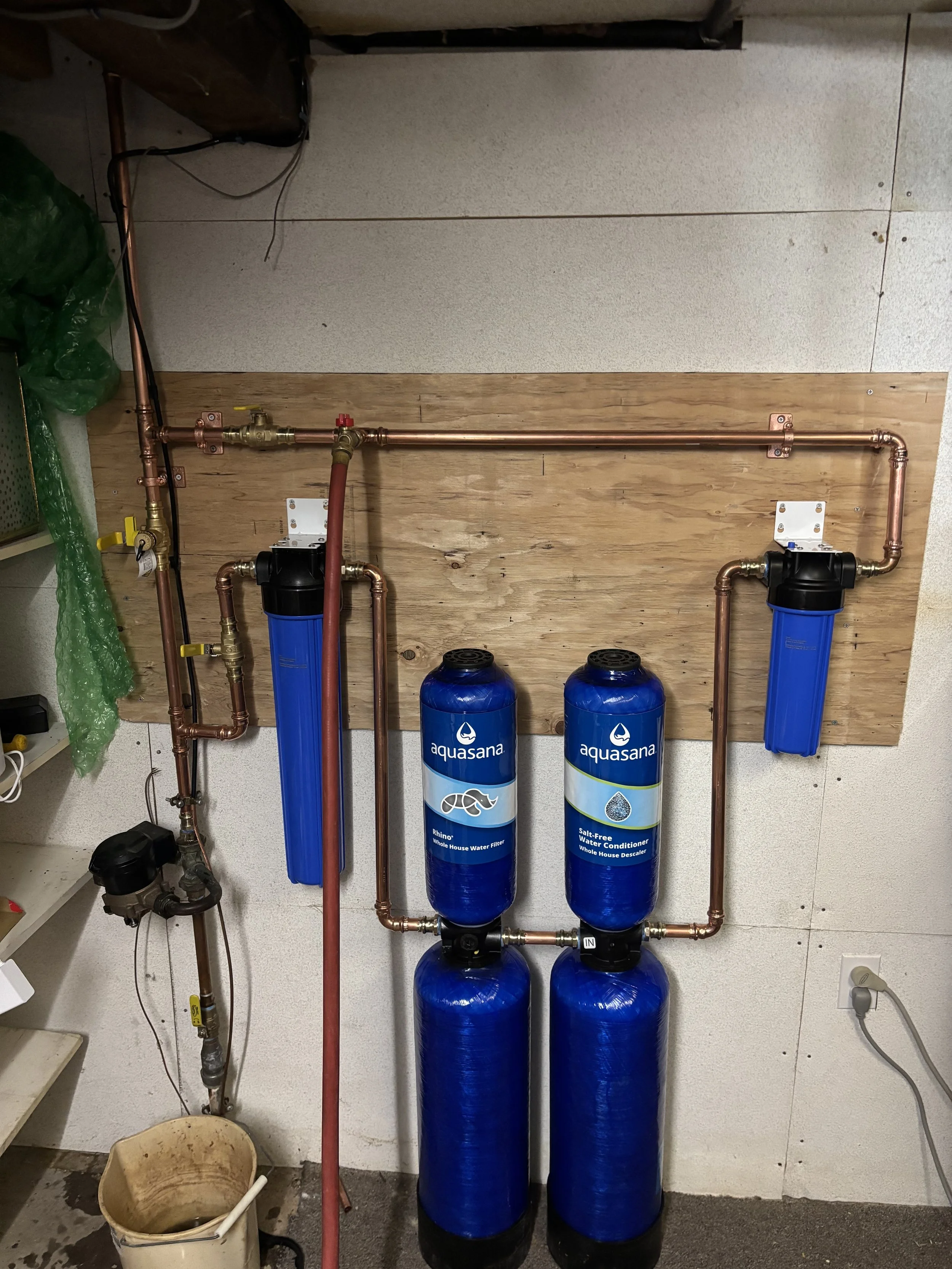 Water filtration system with two large blue Aquasana filters and copper piping on a wooden board in a utility room.