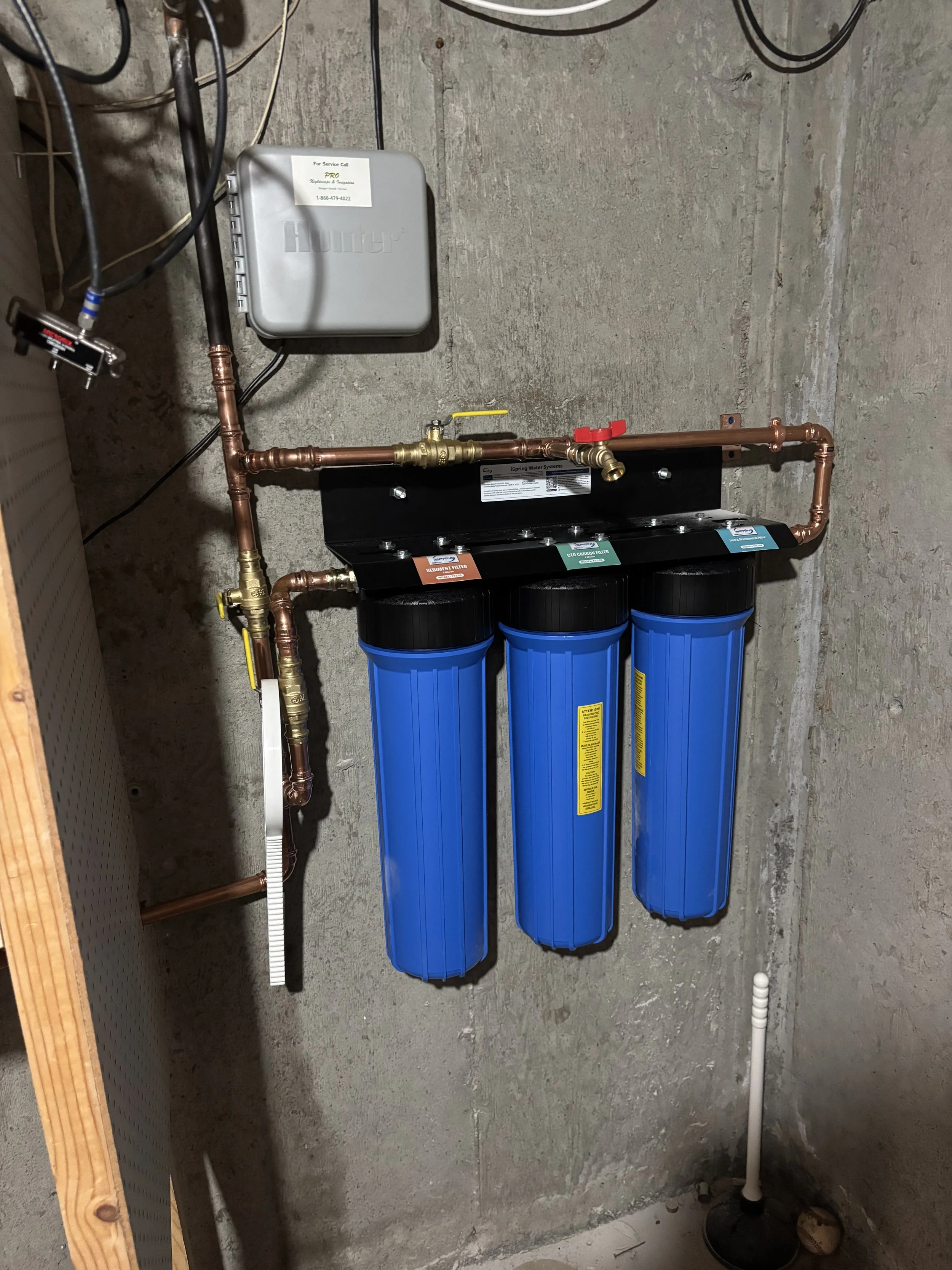 A three-stage water filtration system with blue filter housings, copper piping, and a control valve, installed on a concrete wall.