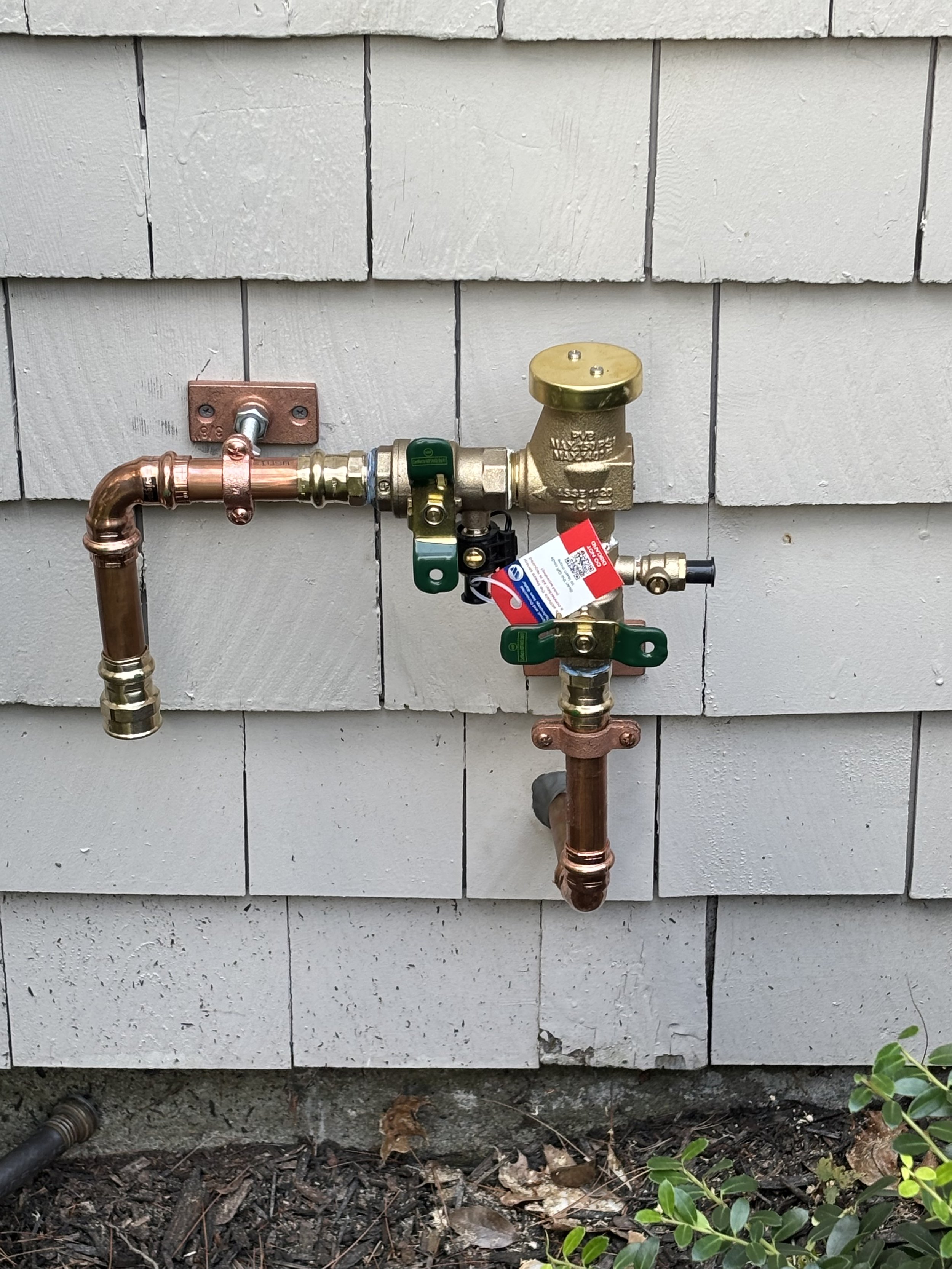 Outdoor plumbing system with copper pipes, a brass valve, and a gauge, mounted on a gray wooden wall.