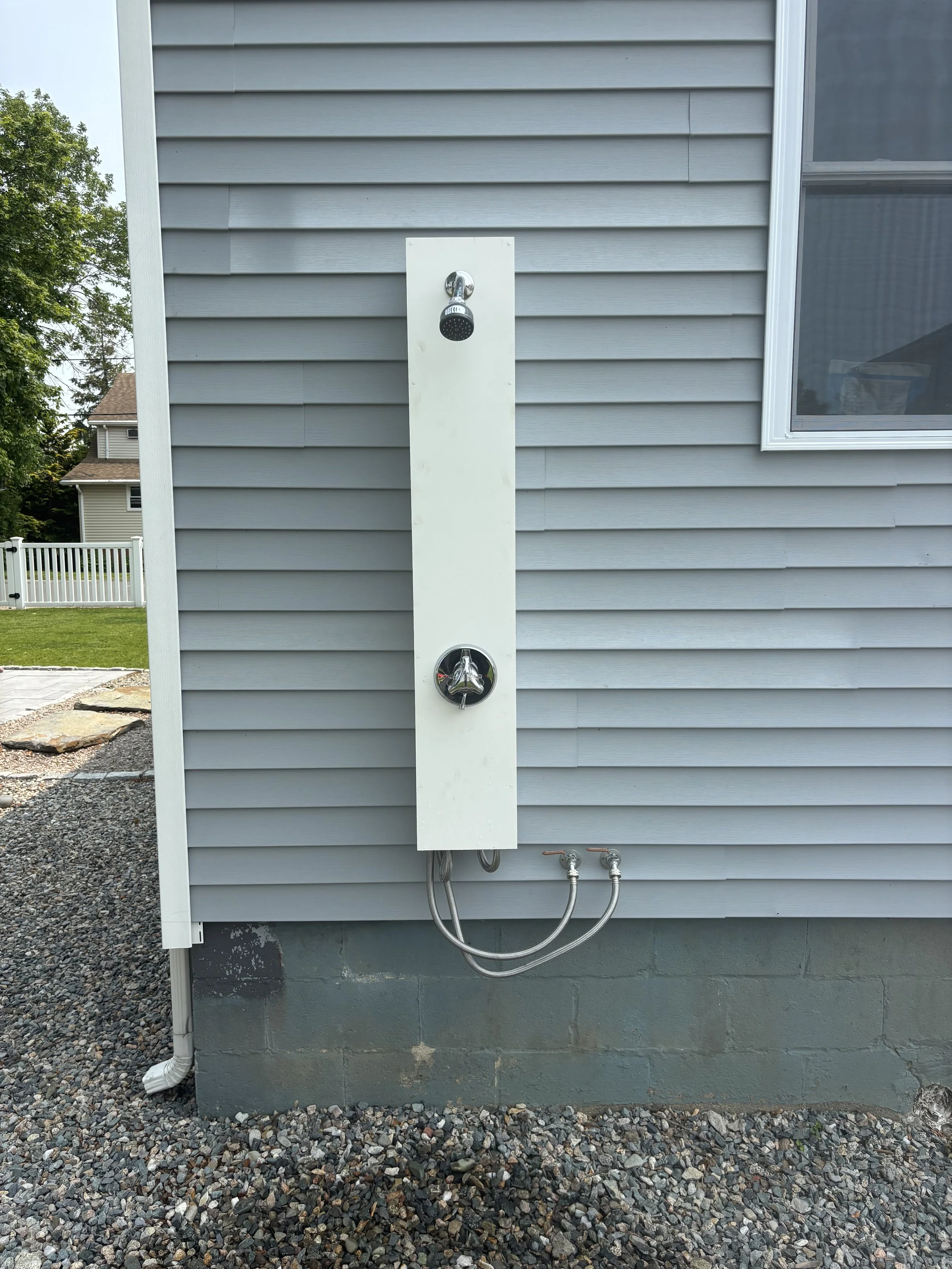 Outdoor shower with gray siding and window, connected to water pipes, installed on the side of a house.
