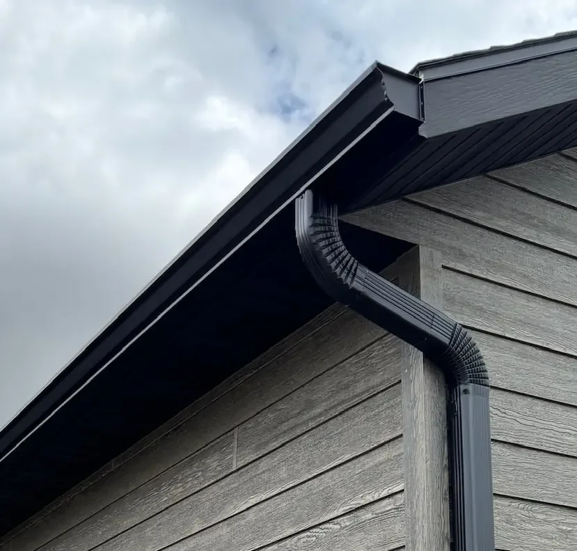 Close-up of a house's roof showing black gutters and downspouts against gray wooden exterior siding and cloudy sky.