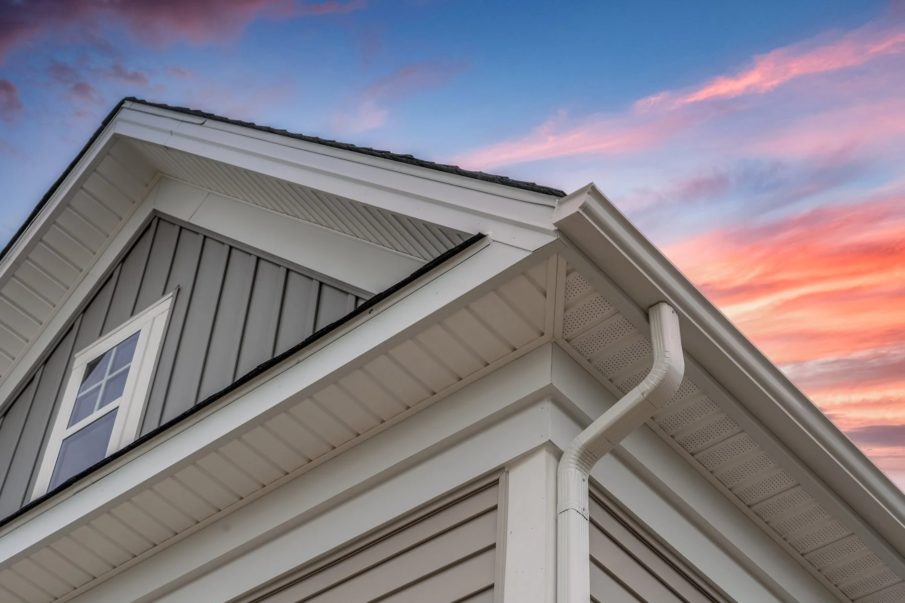 Close-up of the corner of a house with grey vertical siding, white trim, a window, and a gutter system, set against a colorful sunset sky with pink and purple clouds.