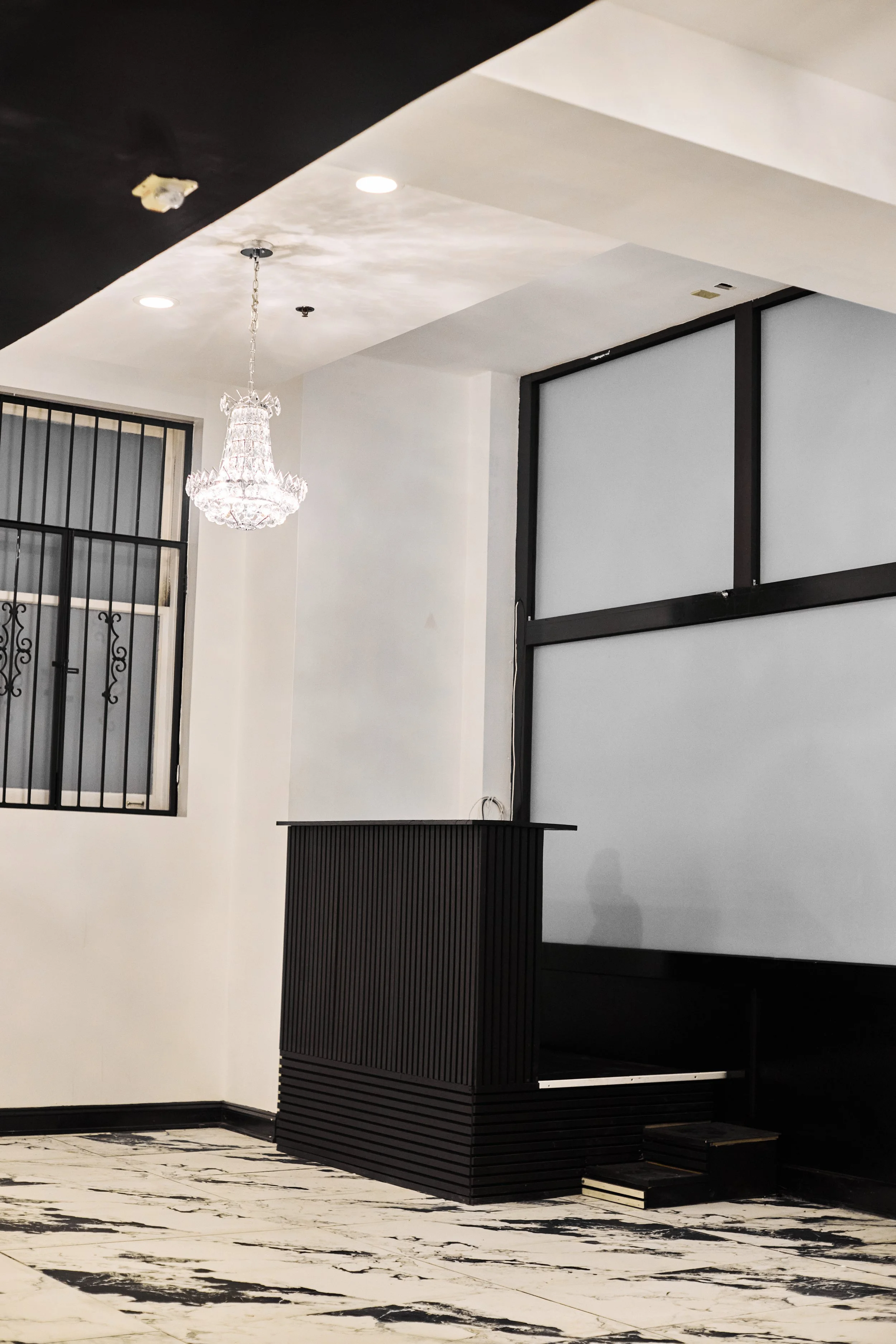 Interior view of a modern space featuring a chandelier, a window with security bars, black and white marble flooring, and black furniture and framing elements.