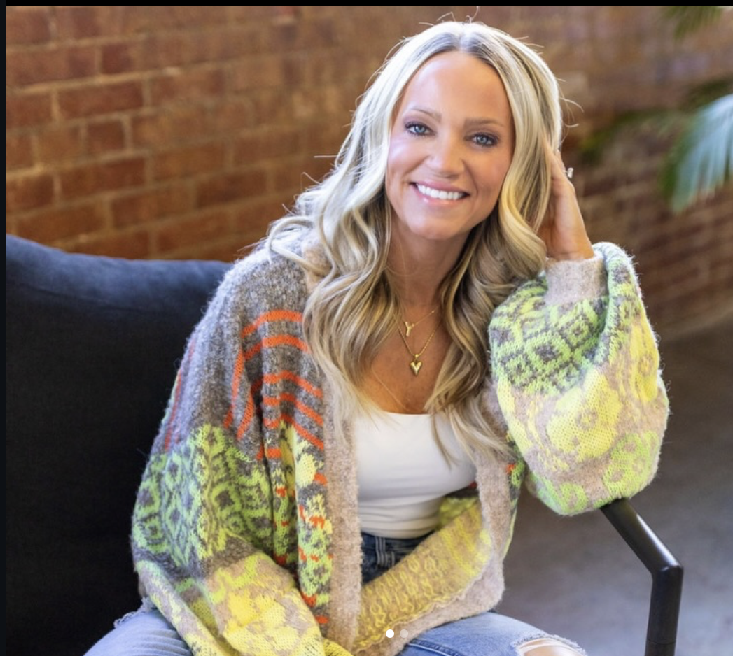 A smiling woman with long, wavy blonde hair, wearing a colorful patterned cardigan over a white shirt, sitting on a black chair in front of a brick wall.