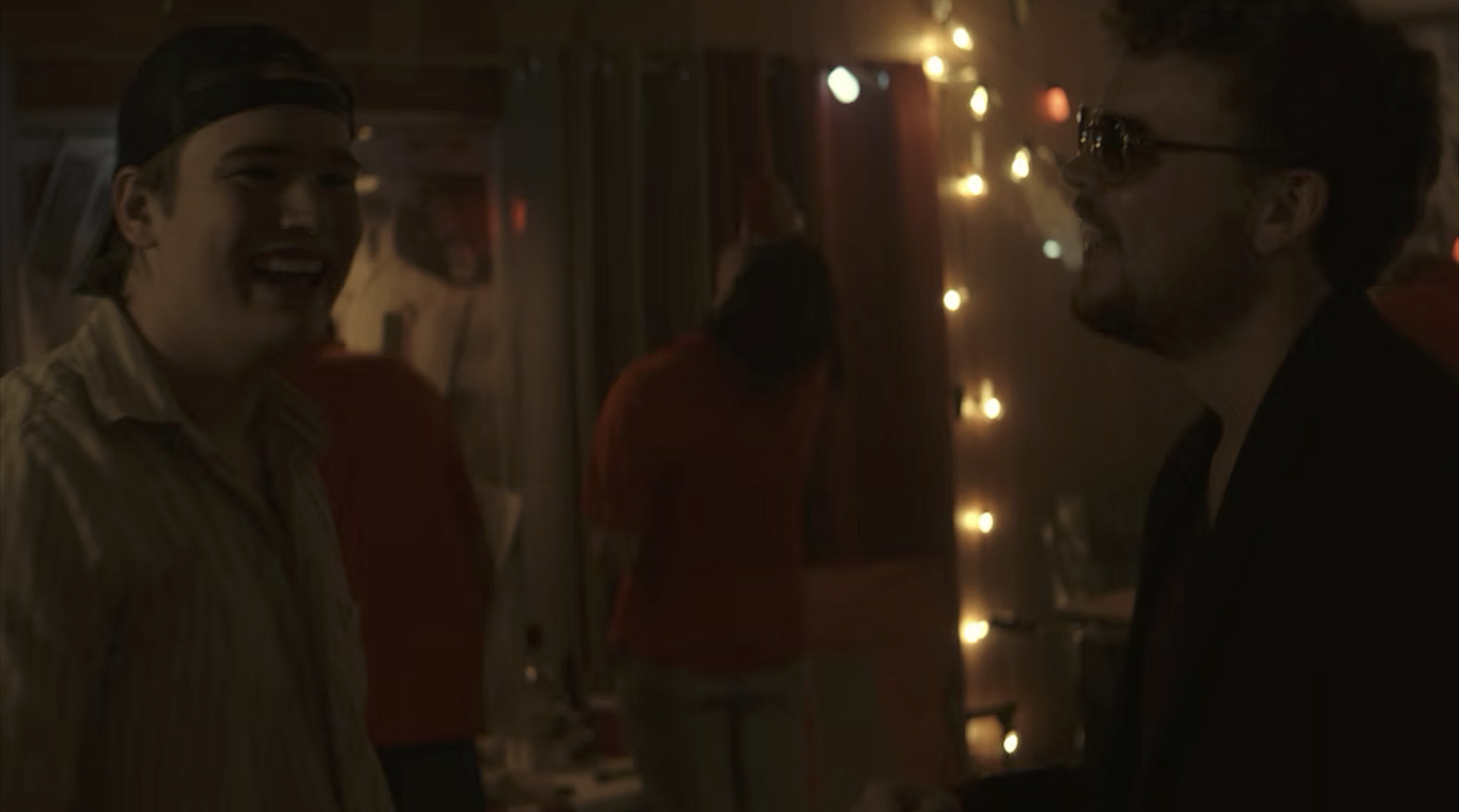 Two men are smiling and talking in a dimly lit room decorated with string lights and posters.