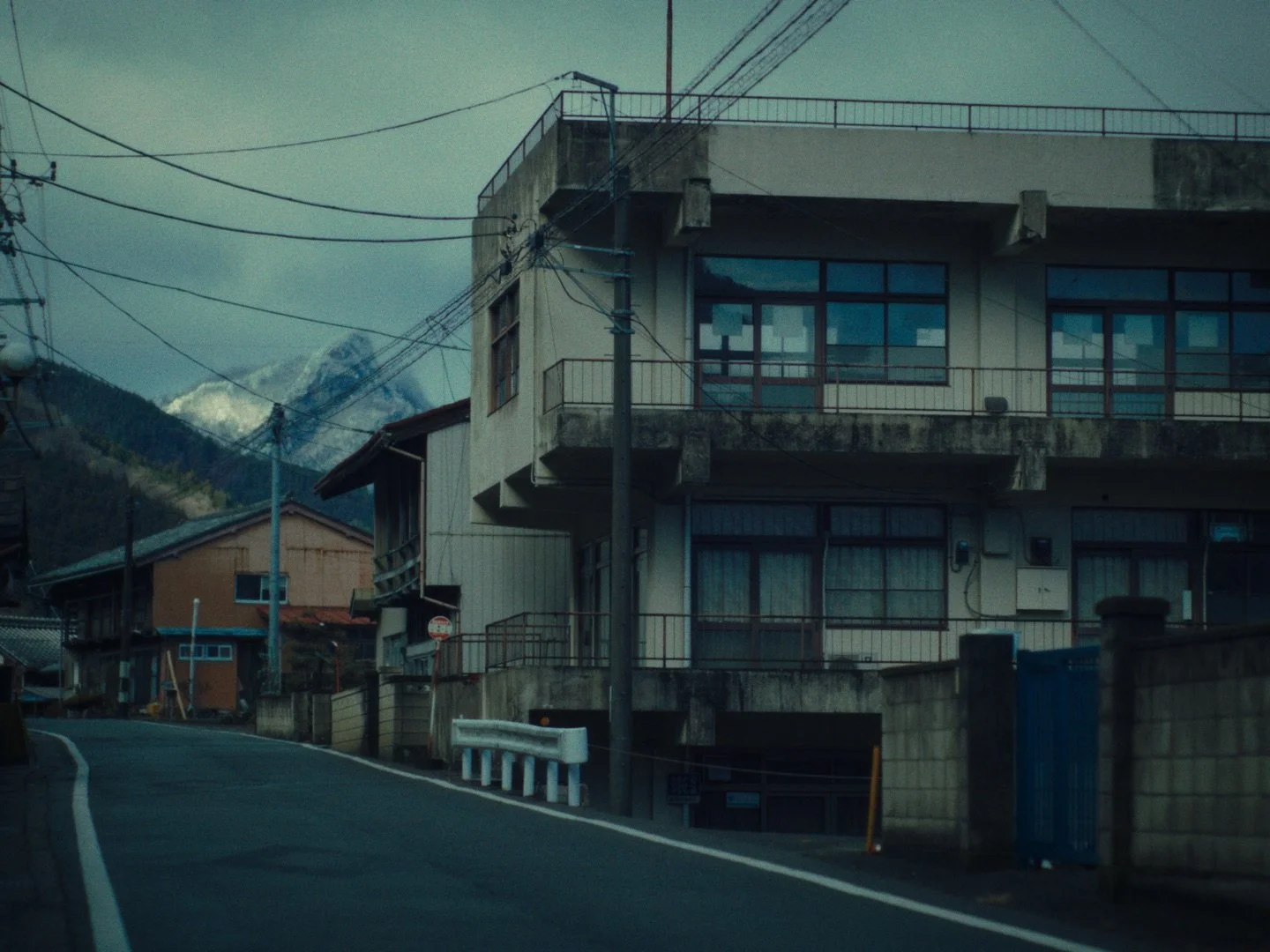 A mountainous residential street scene with multi-story houses, power lines, and snow-capped peaks in the background.