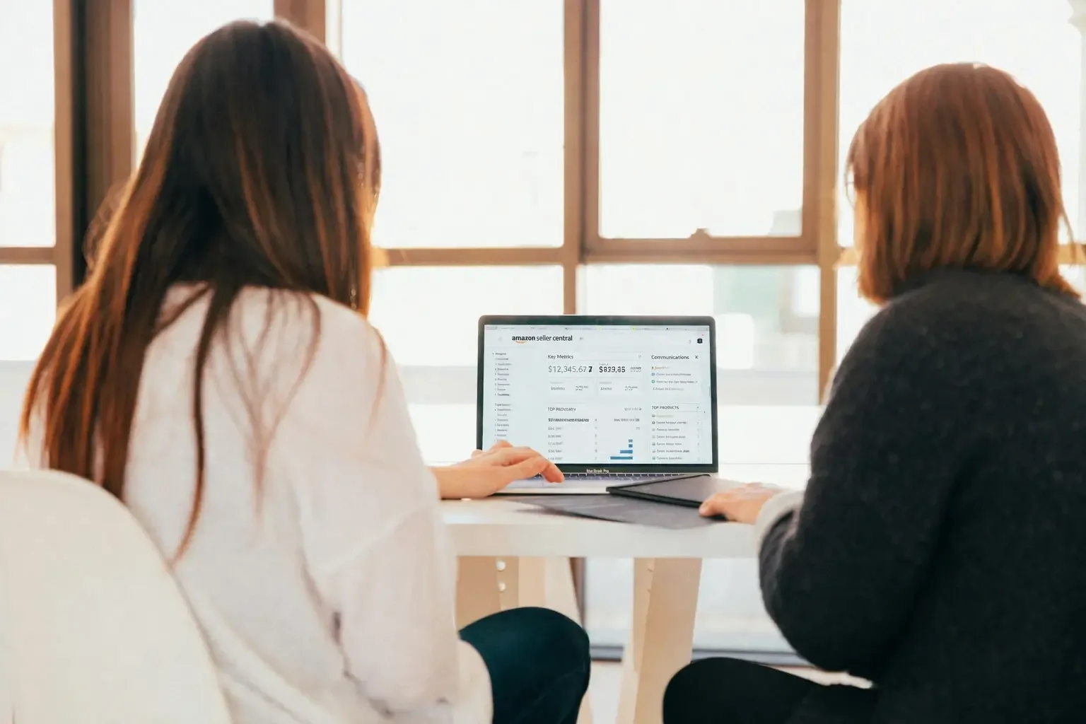 Two women sitting at a table looking at a laptop displaying Amazon seller central dashboard in a bright room with large windows.