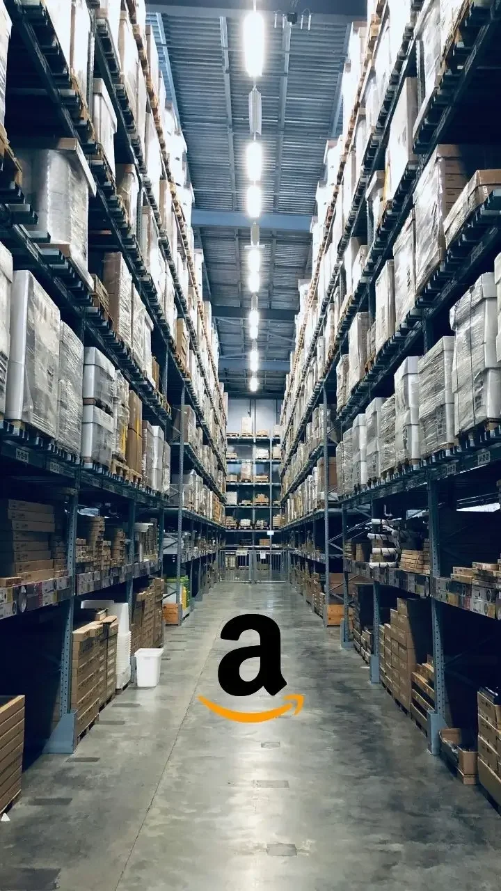 A long aisle in a warehouse with tall shelves stacked with boxes on both sides, lit by overhead fluorescent lights, and with the Amazon icon in the centre.