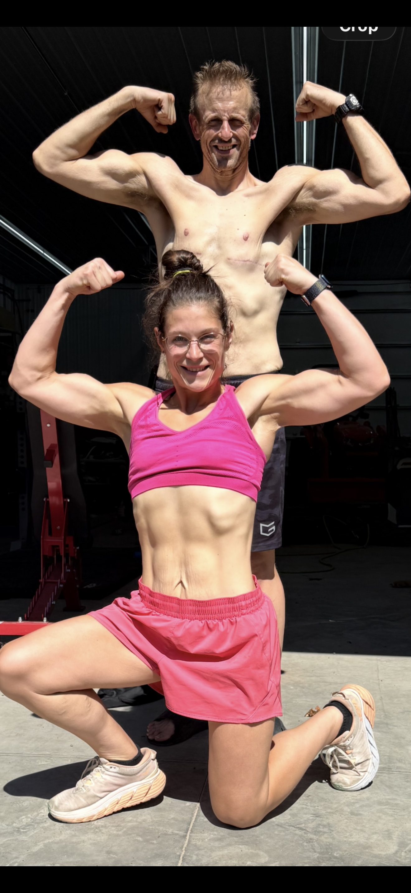 A man and woman posing with flexed muscles in a gym, woman kneeling on one knee, wearing workout clothes, man standing behind her, shirtless, both showing muscular arms and smiling.