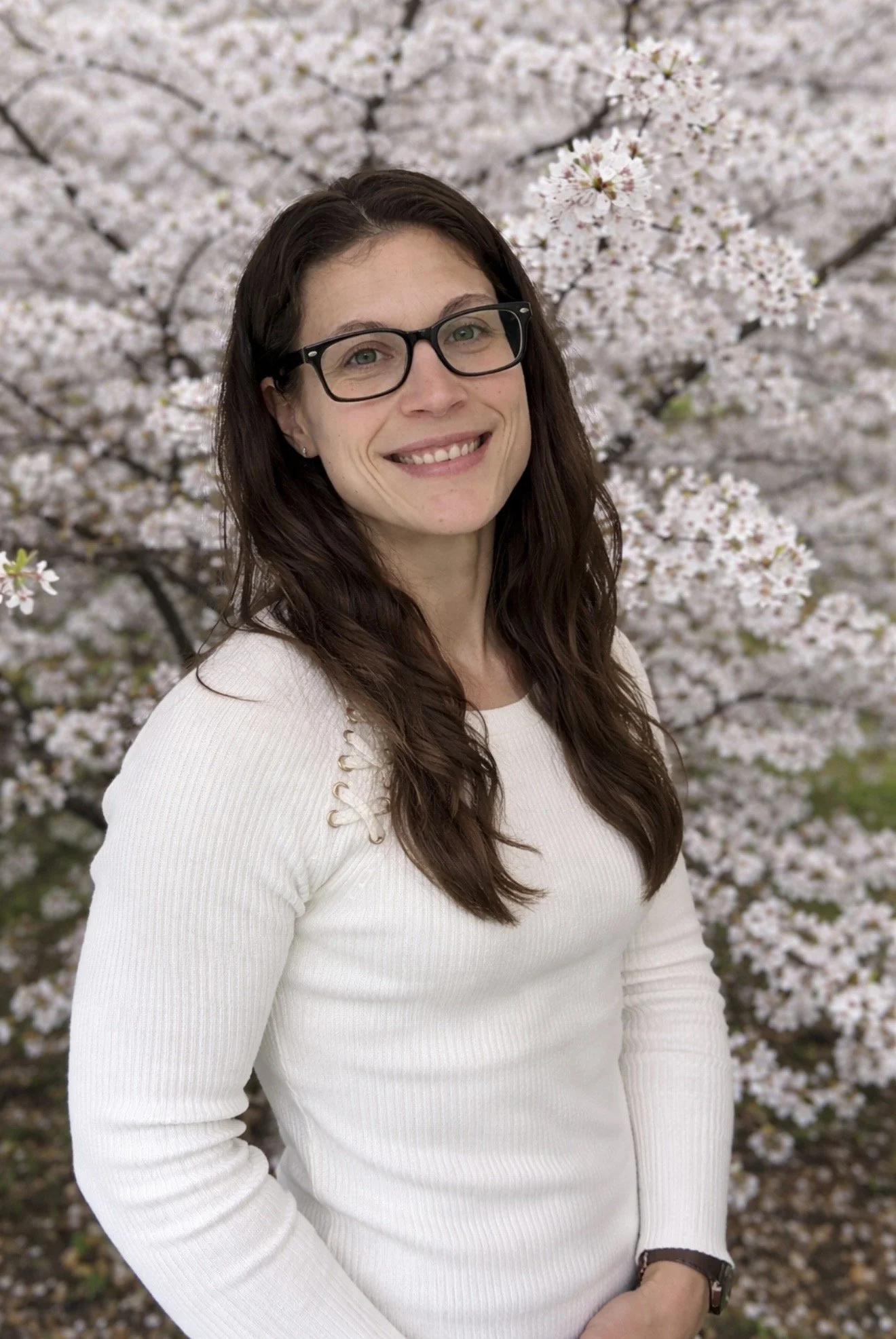A woman with long dark hair and glasses smiling in front of a blooming cherry blossom tree.