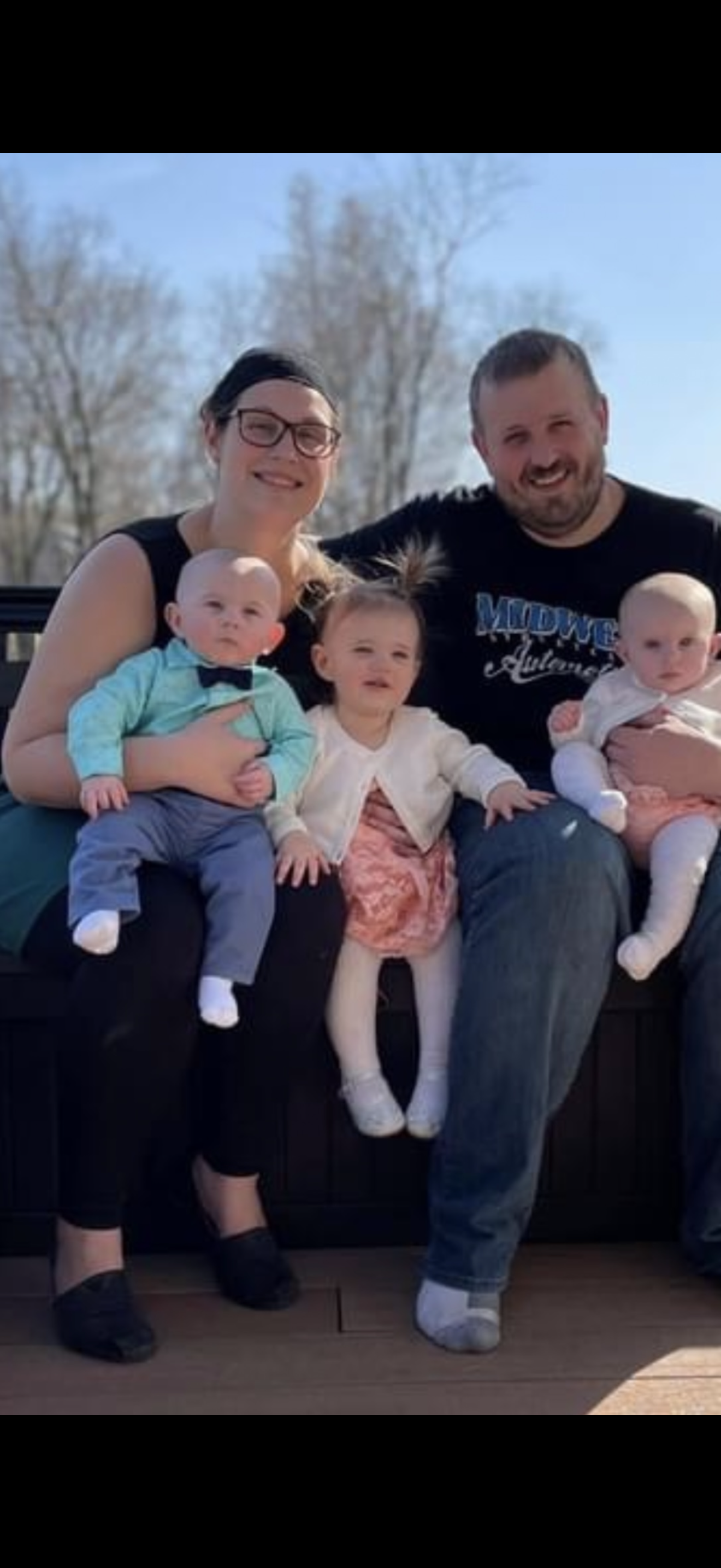 Family photo of two adults and three young children sitting outdoors on a bench, with trees in the background, in daytime.