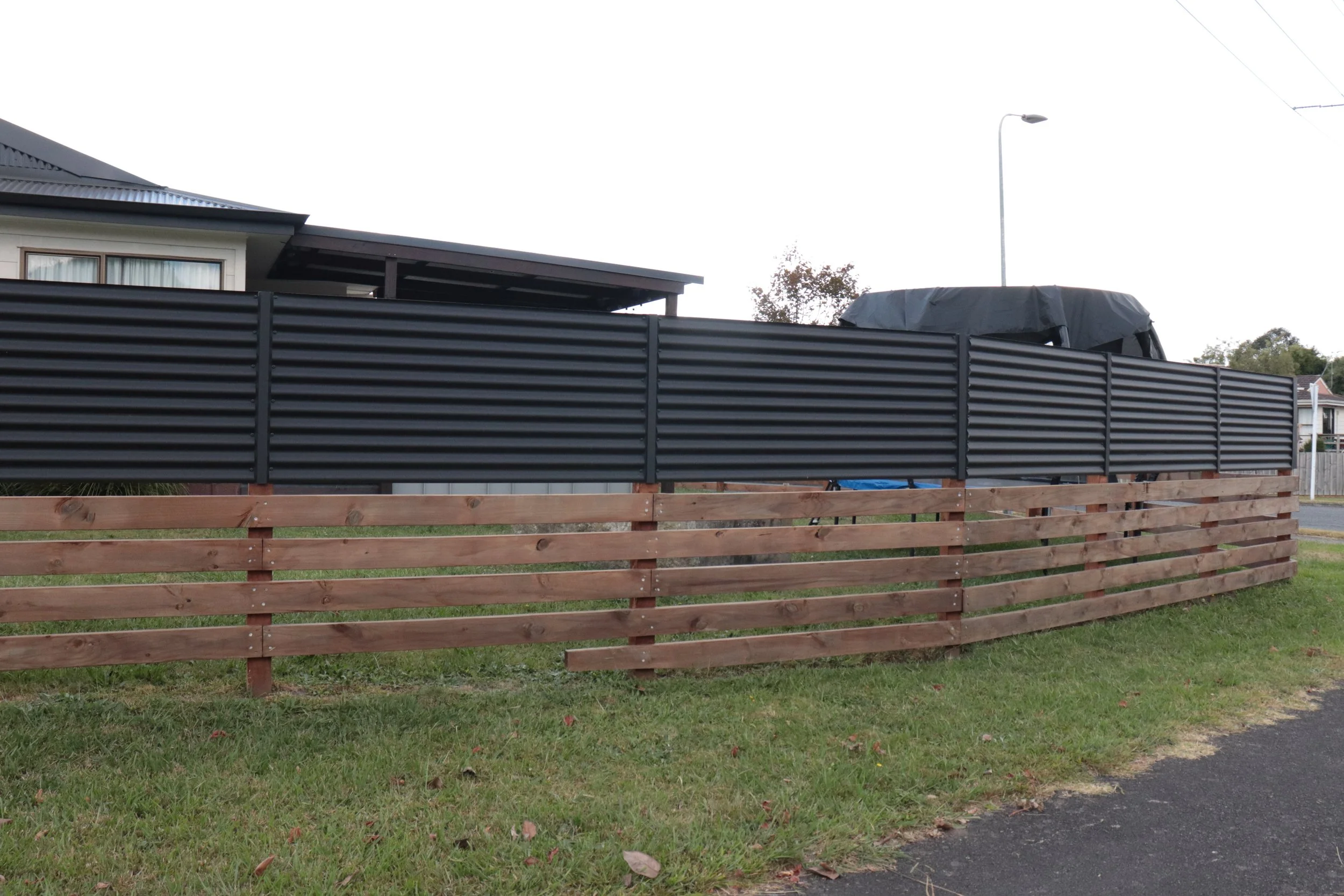 A wooden fence with horizontal planks and black metal panels surrounds a house with a porch and a grey roof. There is a streetlight and trees in the background.