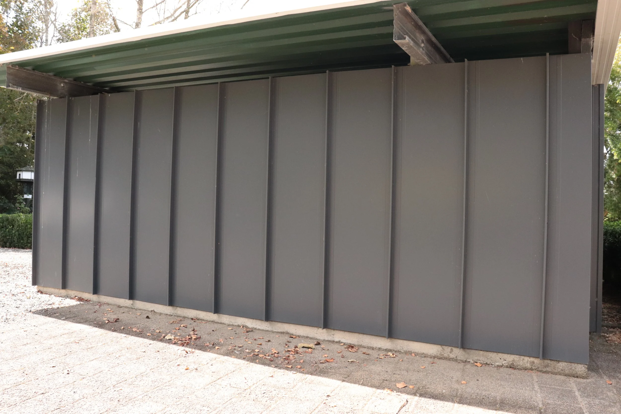 Side of a gray metal building with vertical panels and a green metal roof, located outdoors on a concrete and gravel surface.