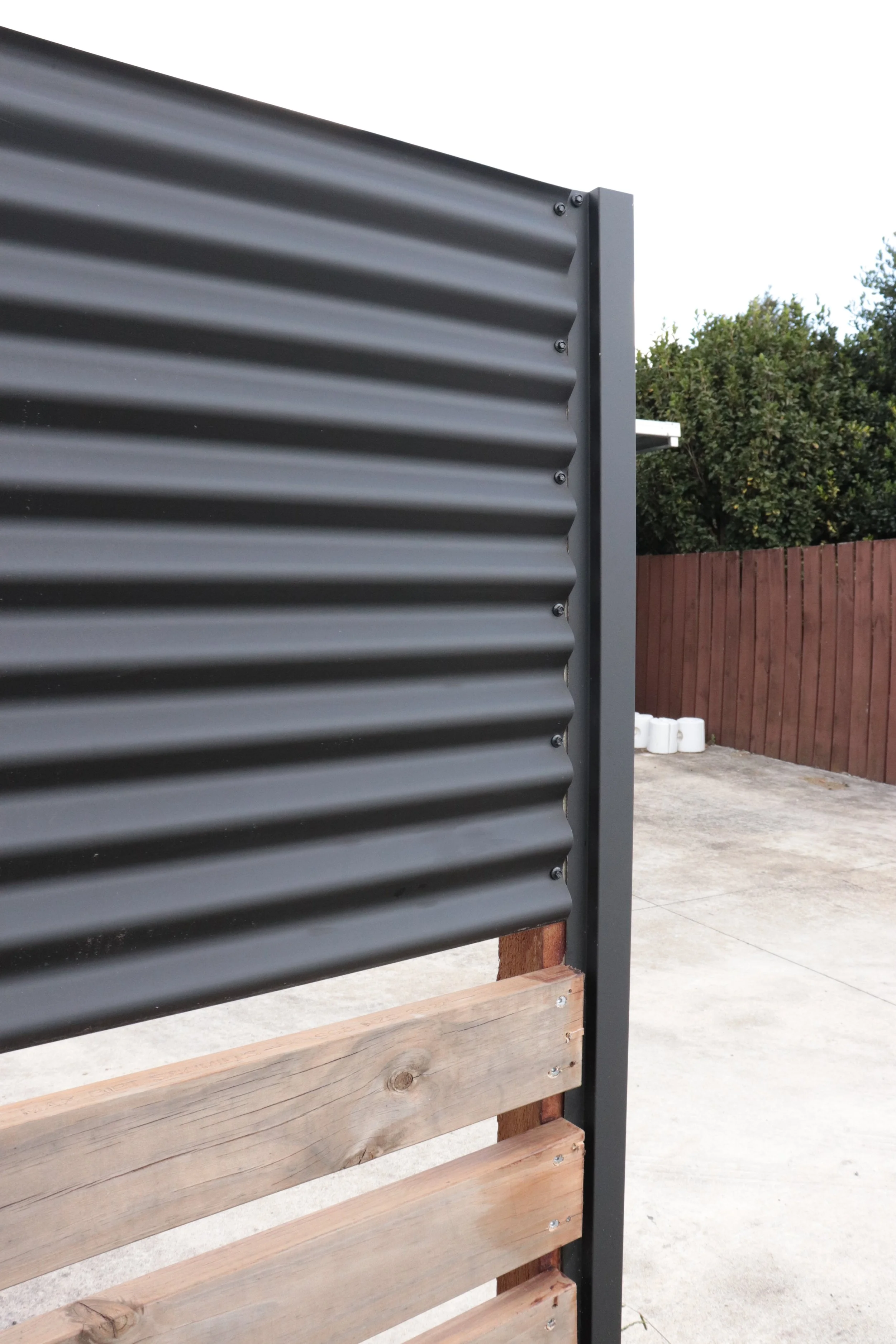 Close-up of a modern outdoor privacy screen with corrugated metal panels and wooden slats, set on a concrete patio with a red wooden fence and greenery in the background.