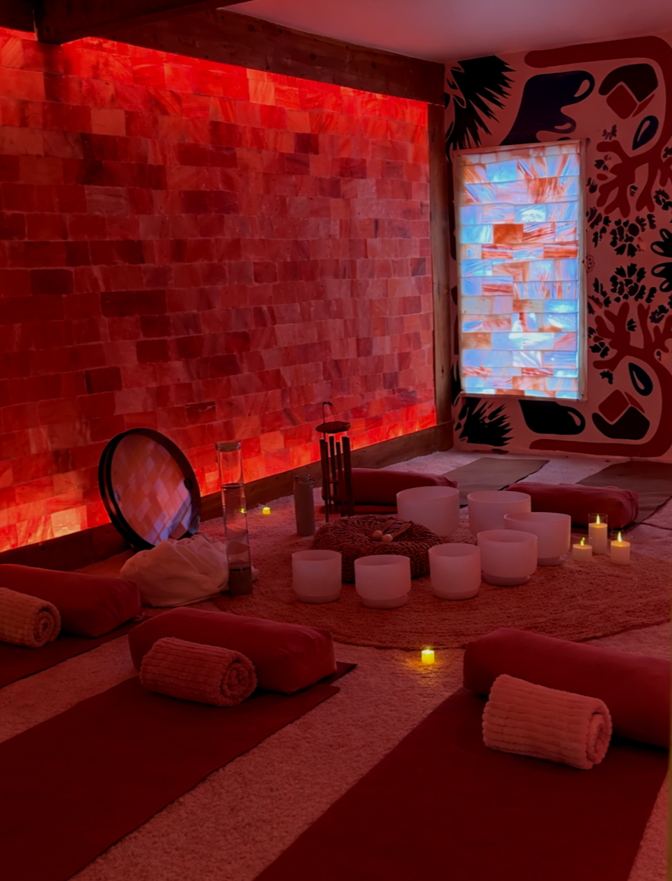 Relaxation room with yoga mats, rolled towels, candles, and singing bowls on a carpeted floor, illuminated by soft red and candlelight, with a salt wall on the left and a colorful window on the right.