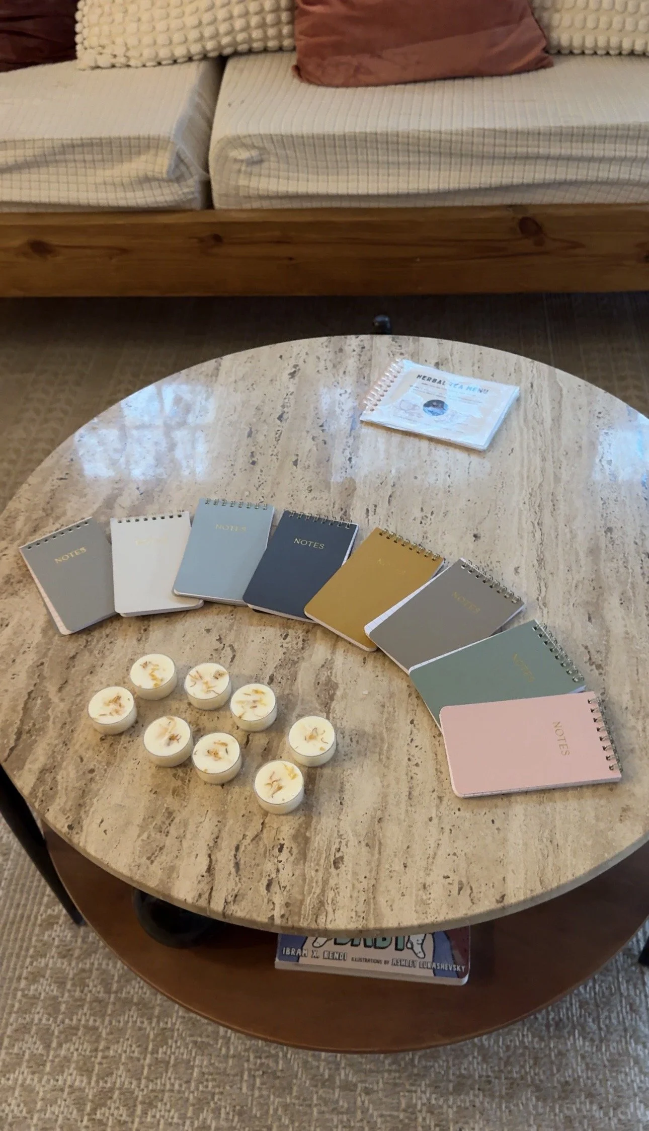 A round marble-top table with paper notebooks in pastel colors, six white tea light candles, and a pamphlet titled 'Herbal Tea Menu' on top of the table. A lower shelf holds some magazines and books. In the background, a beige bed with checkered bedd