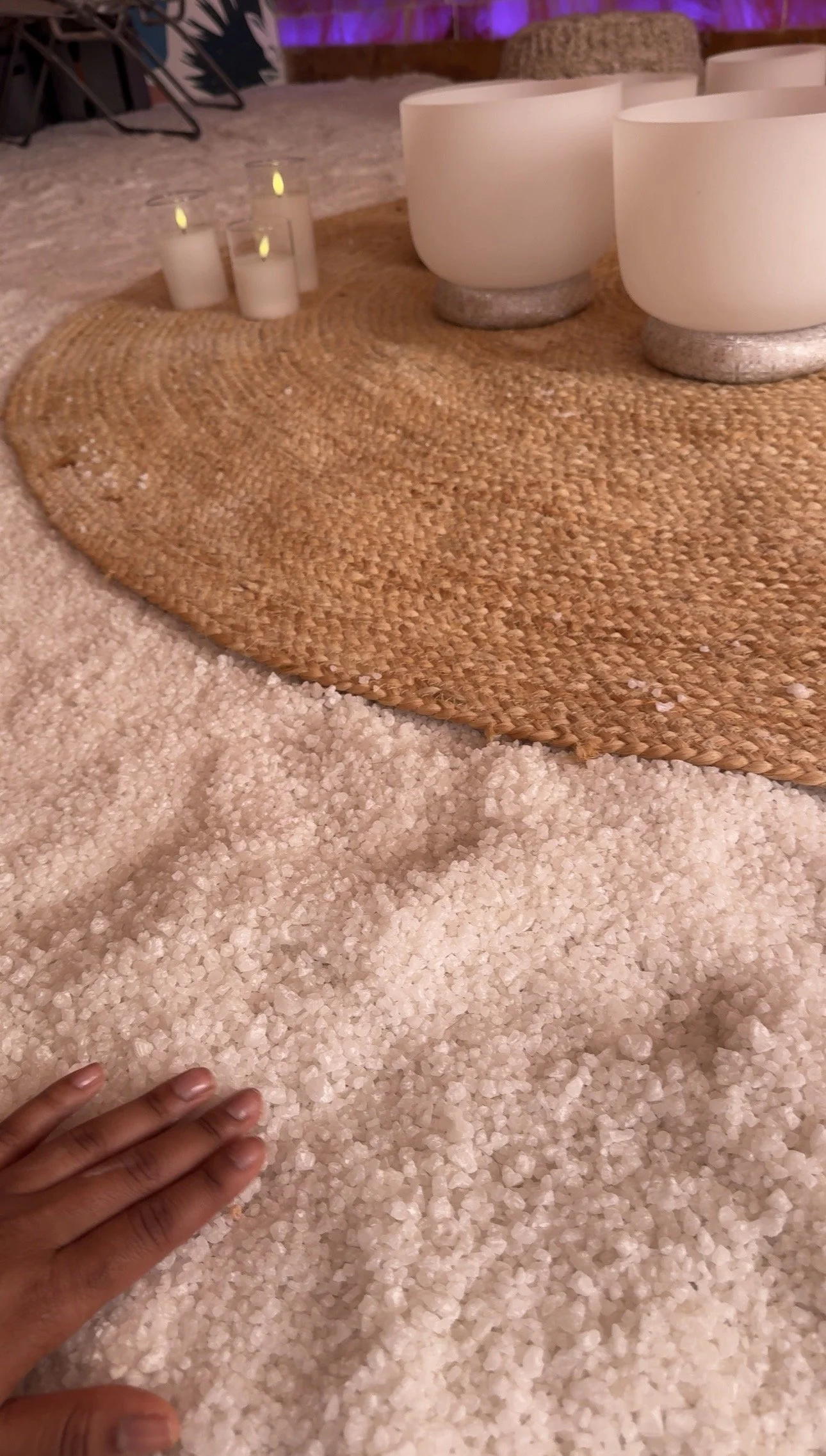 Close-up of a cozy interior with a woven circular rug on top of a plush white carpet, white modern bowls on a table, and candle decorations in the background.