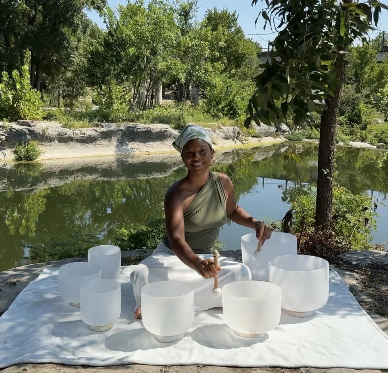 A woman playing crystal singing bowls in Austin Tx outdoors near a pond, surrounded by trees and rocks, with sunlight shining on her and the bowls.