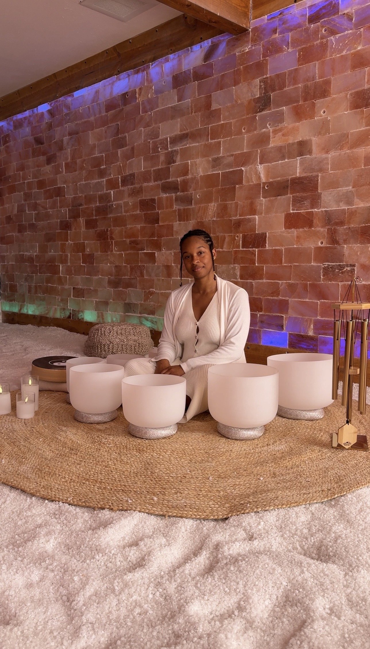 A woman sitting with singing bowls and candles on a woven rug against a brick wall with blue and purple lighting.