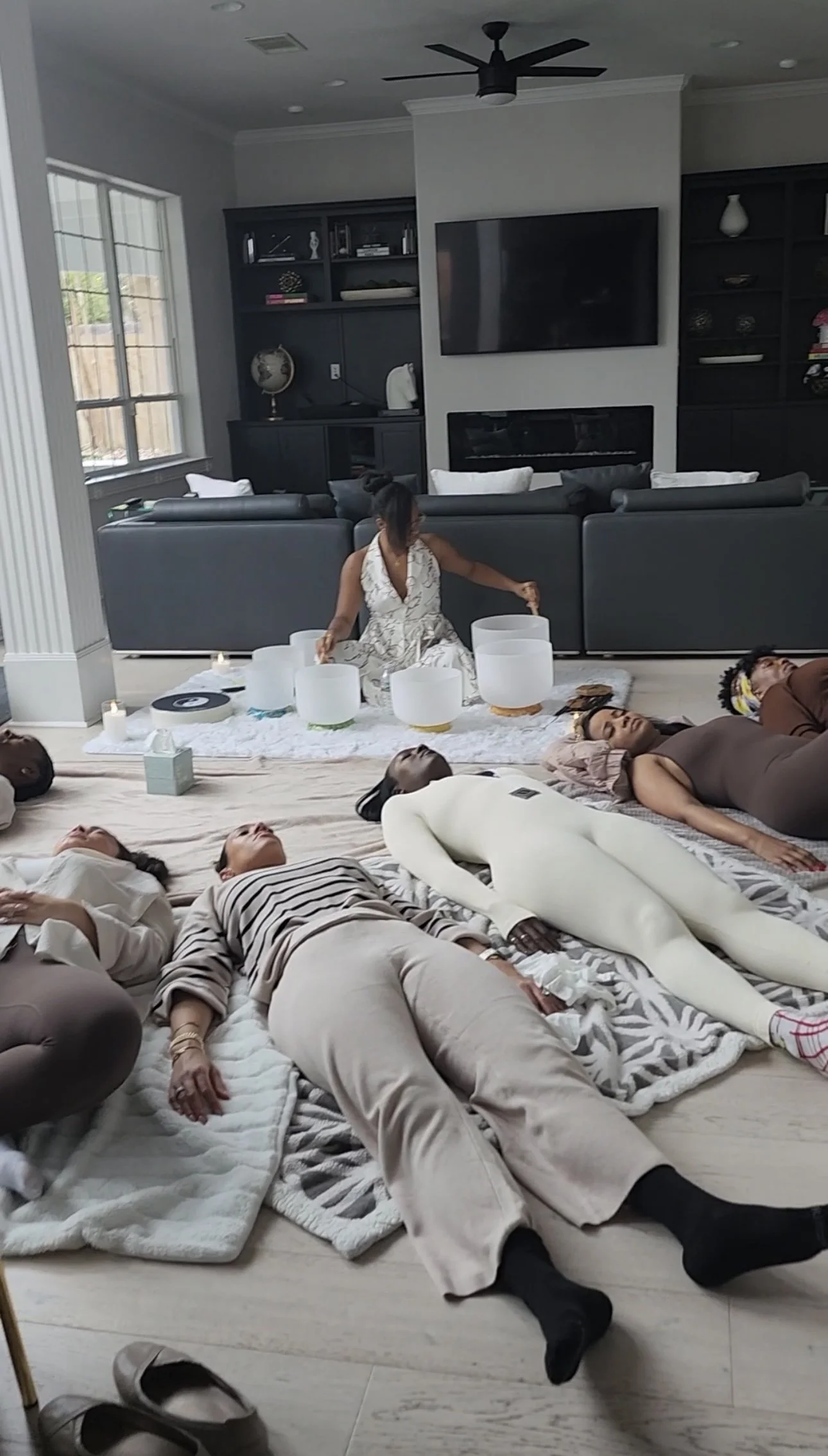 Group of people lying on blankets on the floor with a woman sitting cross-legged in the background, playing crystal sound bowls in a living room for a group sound bath in Austin, Tx.