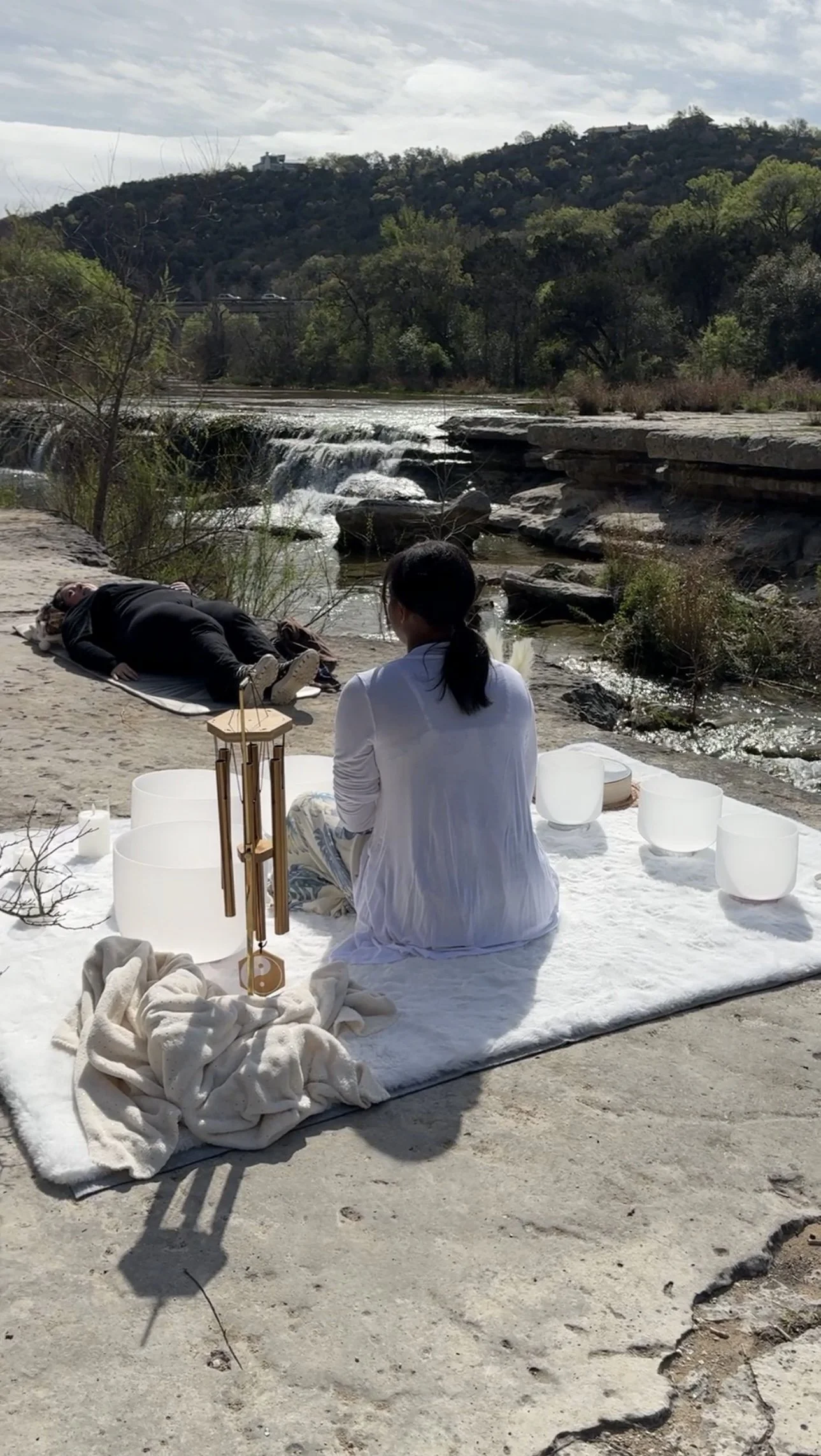 A woman in white robes sitting on a blanket near a river with water bowls around her, while a person lies on a mat by the riverbank. The setting is outdoors with a scenic view of trees, rocks, and a small waterfall in the background.