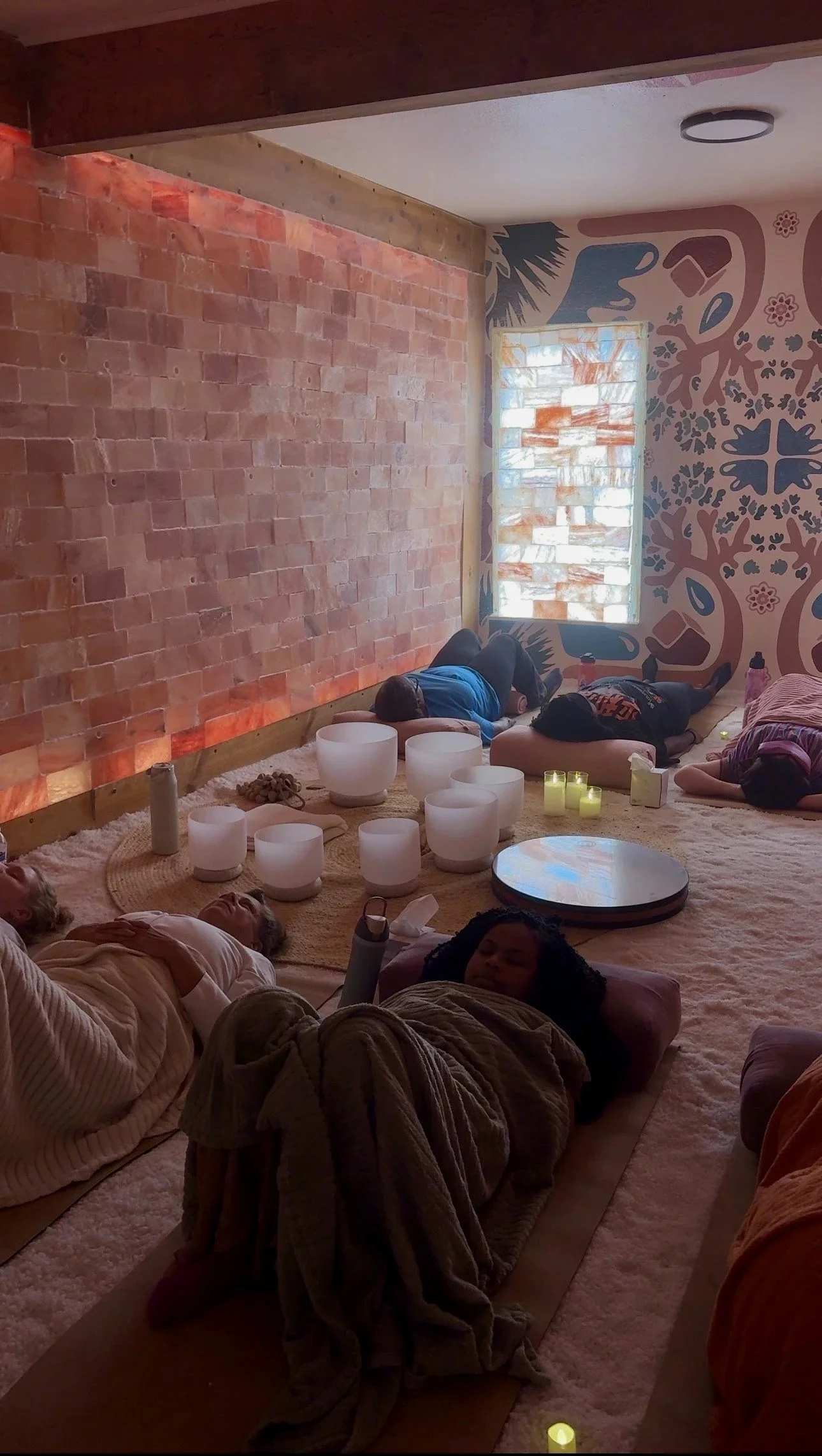 People lying down on mats in a room with a brick wall, a window with salt lamp tiles, and a wall mural with floral and abstract designs. Several white singing bowls and candles are on the floor.