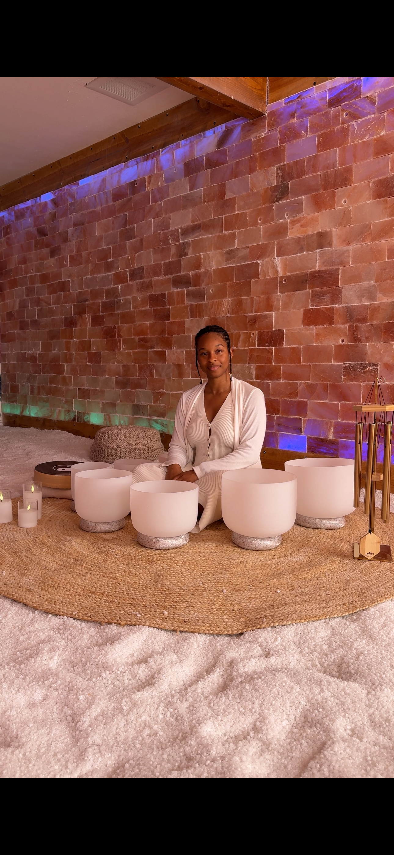 A woman sitting on a woven rug surrounded by white singing bowls, candles, and wind chimes against a red brick wall with blue lighting accents.