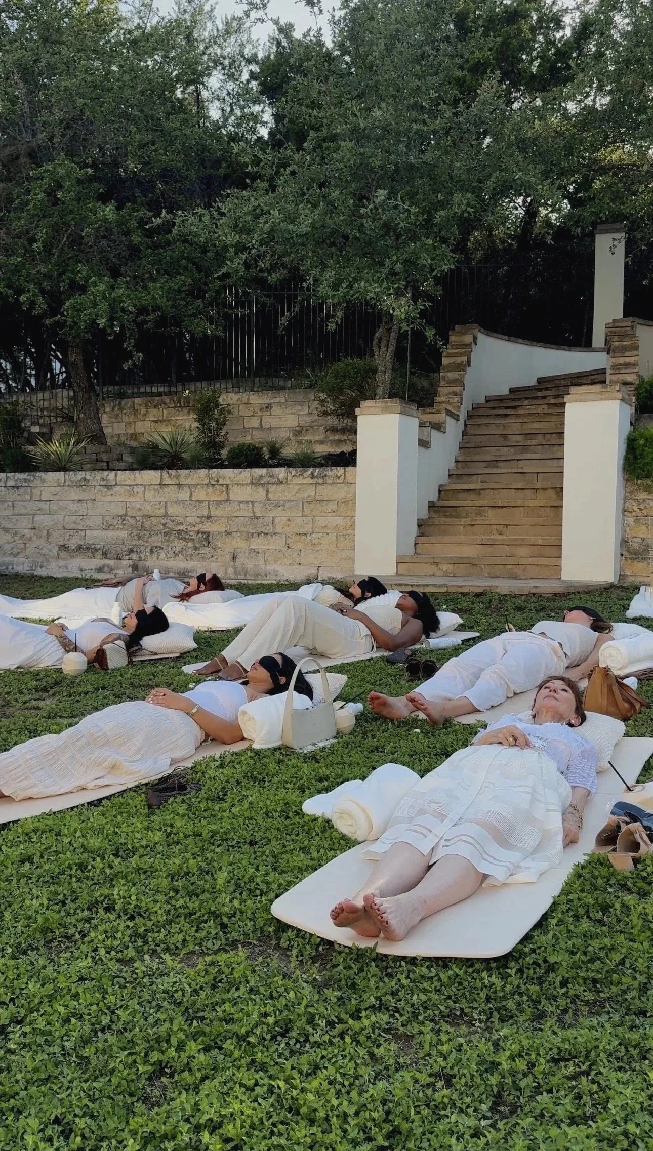 People lying on white mats and blankets on green grass, some with eye masks, in an outdoor garden area with stone stairs and trees.