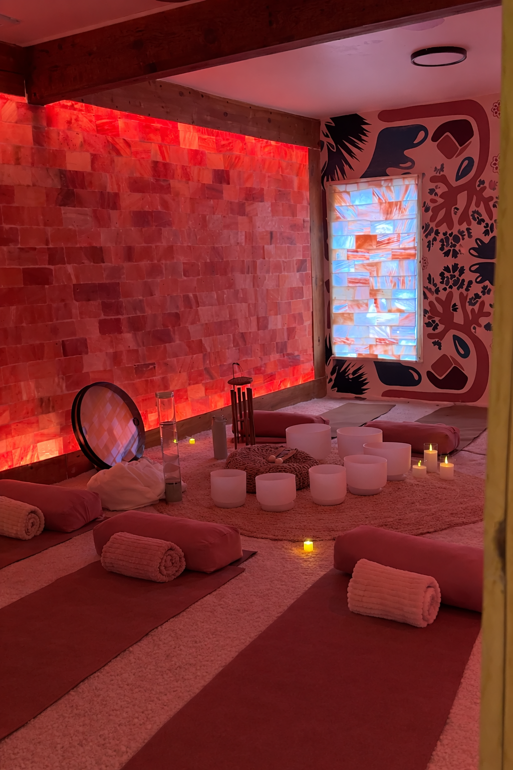 A cozy room with pink and red lighting, decorated with a textured brick wall and black and pink floral mural. There are yoga mats, rolled towels, candles, and white bowls arranged on a circular rug, creating a calming atmosphere.