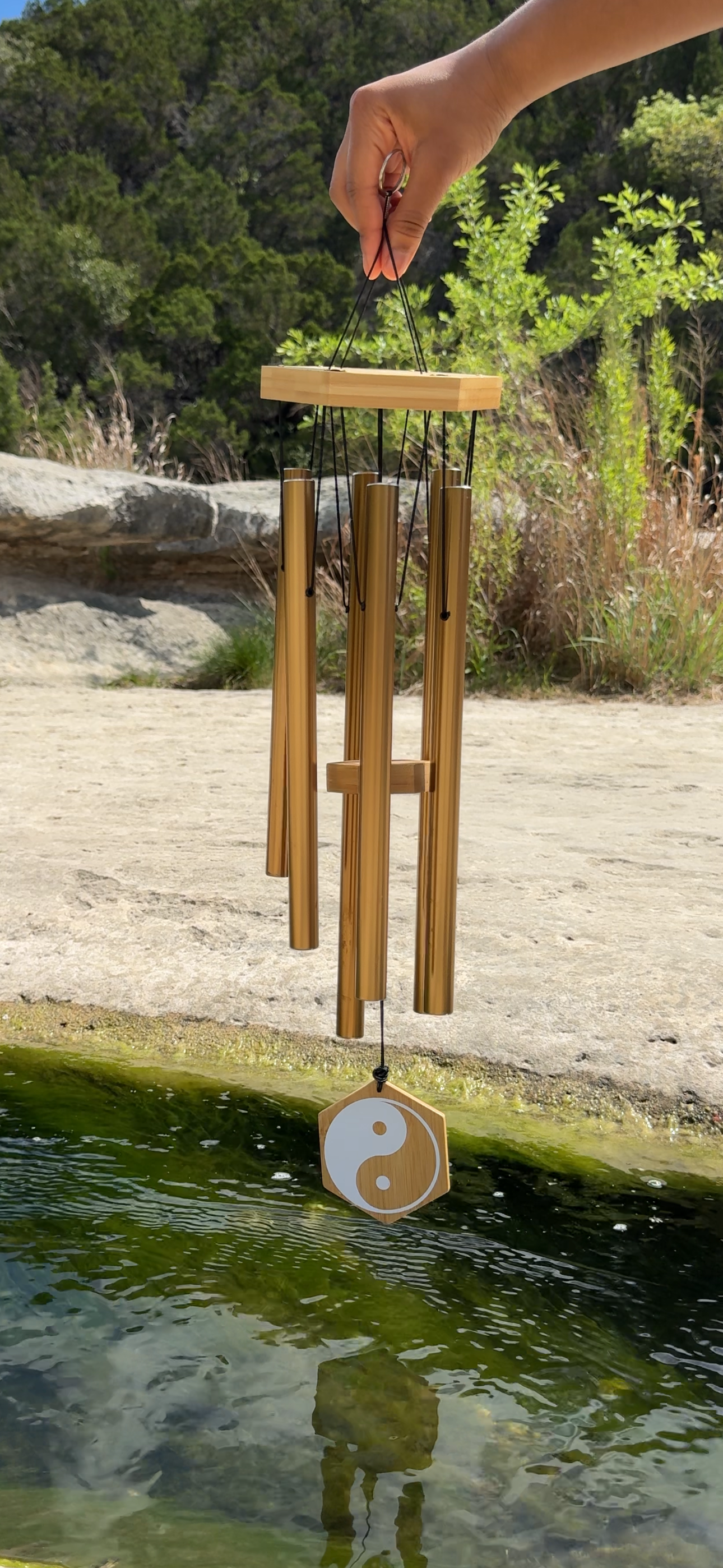 Person holding a wooden wind chime with a yin-yang symbol in front of a natural outdoor setting with rocks, trees, and a body of water.