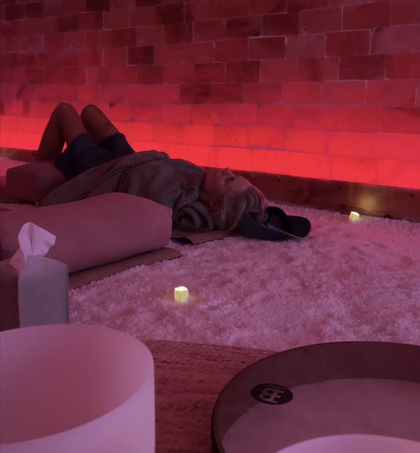 A person lying on the floor in a cozy room with red lighting, a pillow, a blanket, and a pair of headphones nearby, surrounded by small candles on a pink fluffy rug.