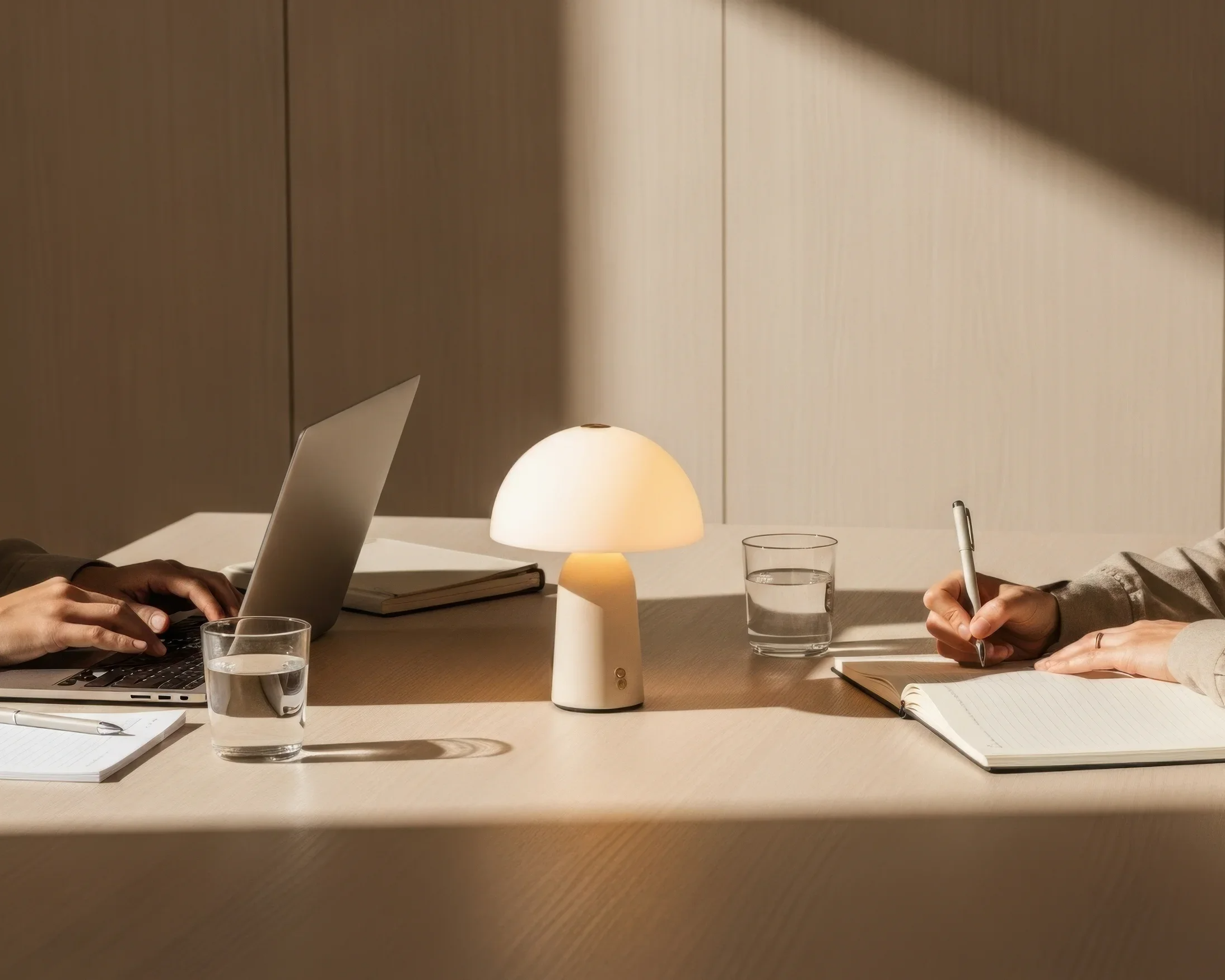 Two people sitting at a wooden table with notebooks, glasses of water, a lamp, and a laptop, engaged in work or discussion.