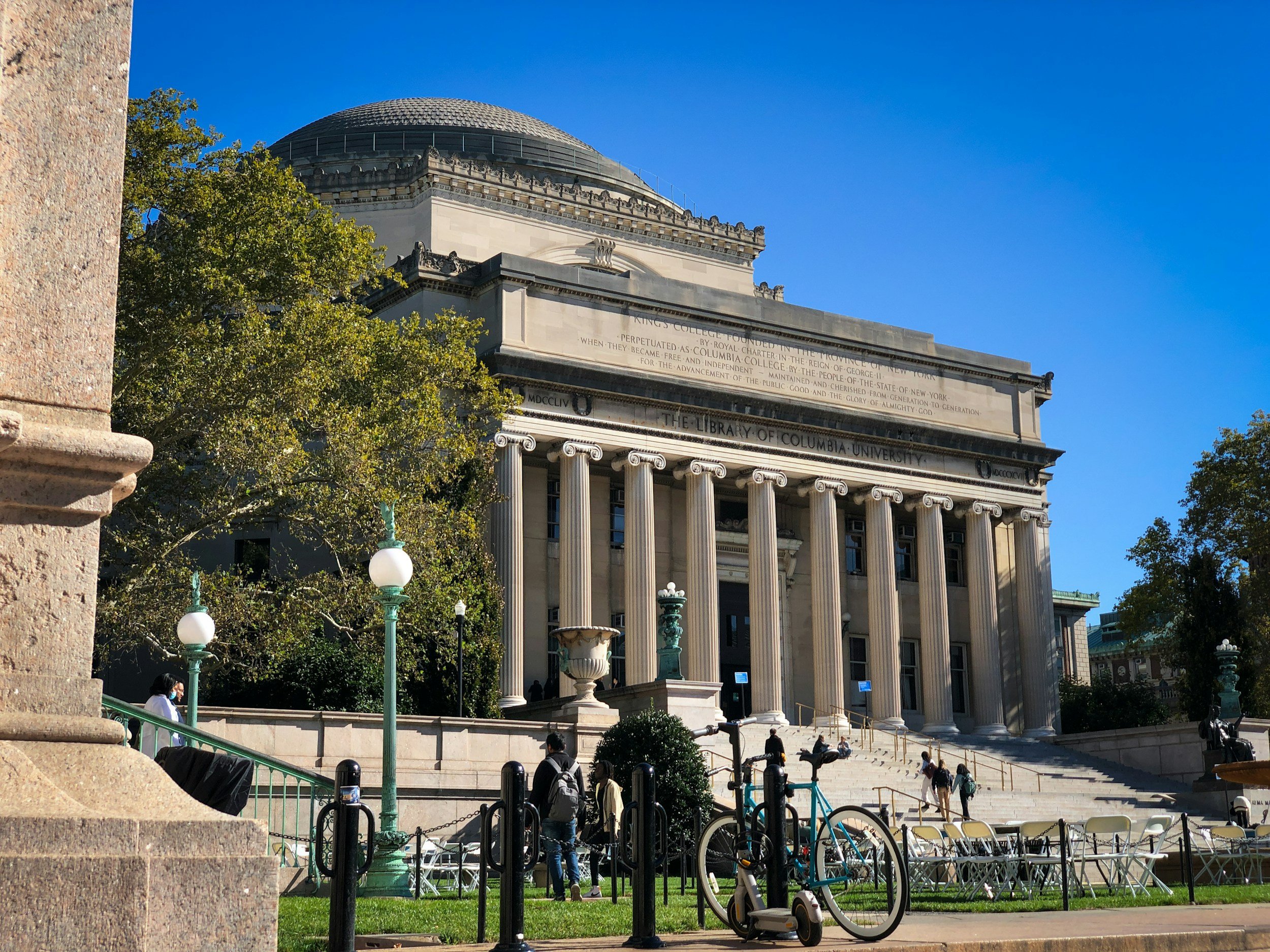 The Library of Columbia University building with classical columns, stairs, and trees in front, under a clear blue sky.