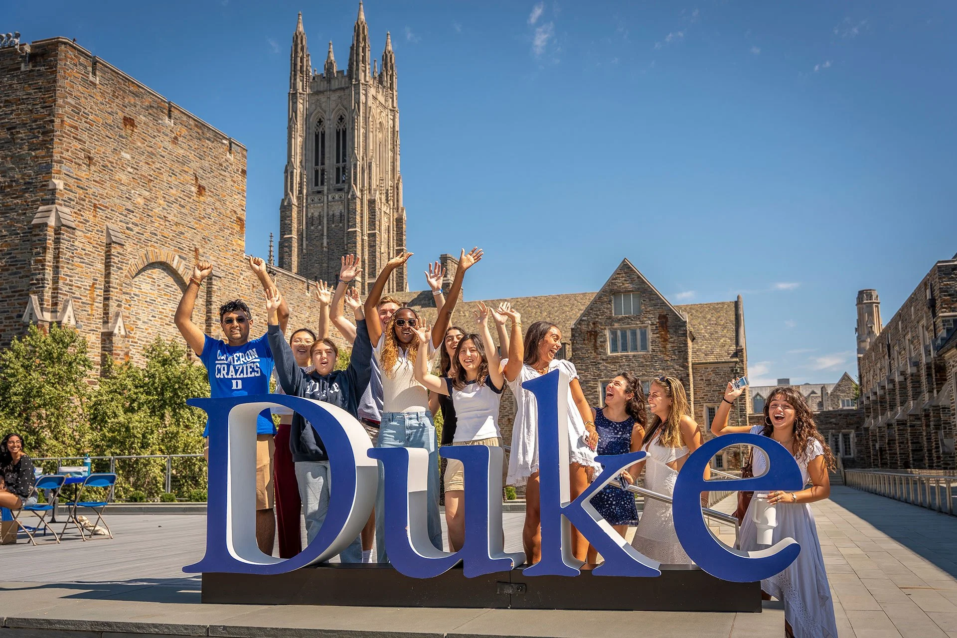 A group of diverse students celebrating behind a large blue and white 'Duke' sign on a university campus with historic stone buildings and a tall spire in the background on a sunny day.